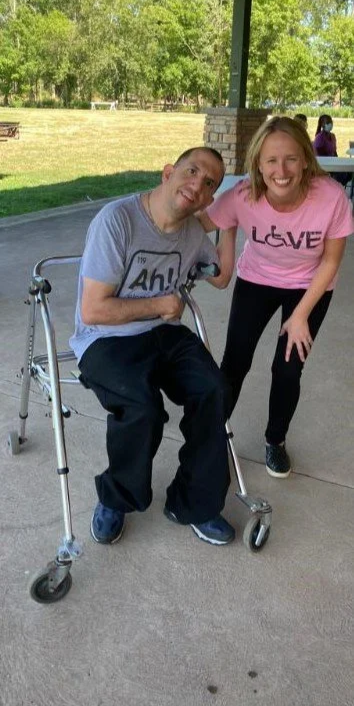 Two people, a man using a walker and a woman, smiling outdoors under a pavilion, with trees and a grassy field in the background.
