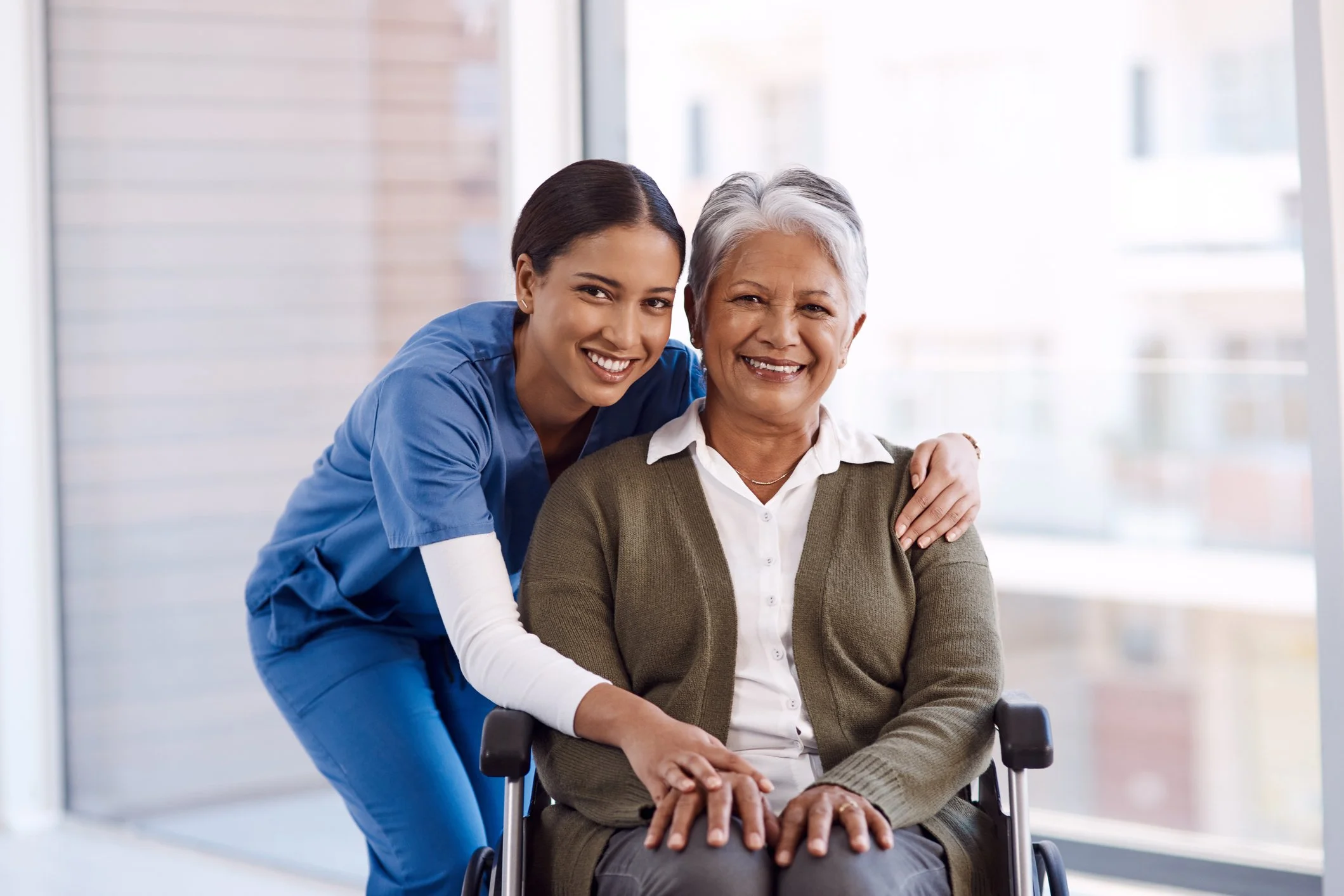 A young female caregiver in blue scrubs smiling with an elderly woman in a wheelchair, both smiling and looking at the camera in a bright room.