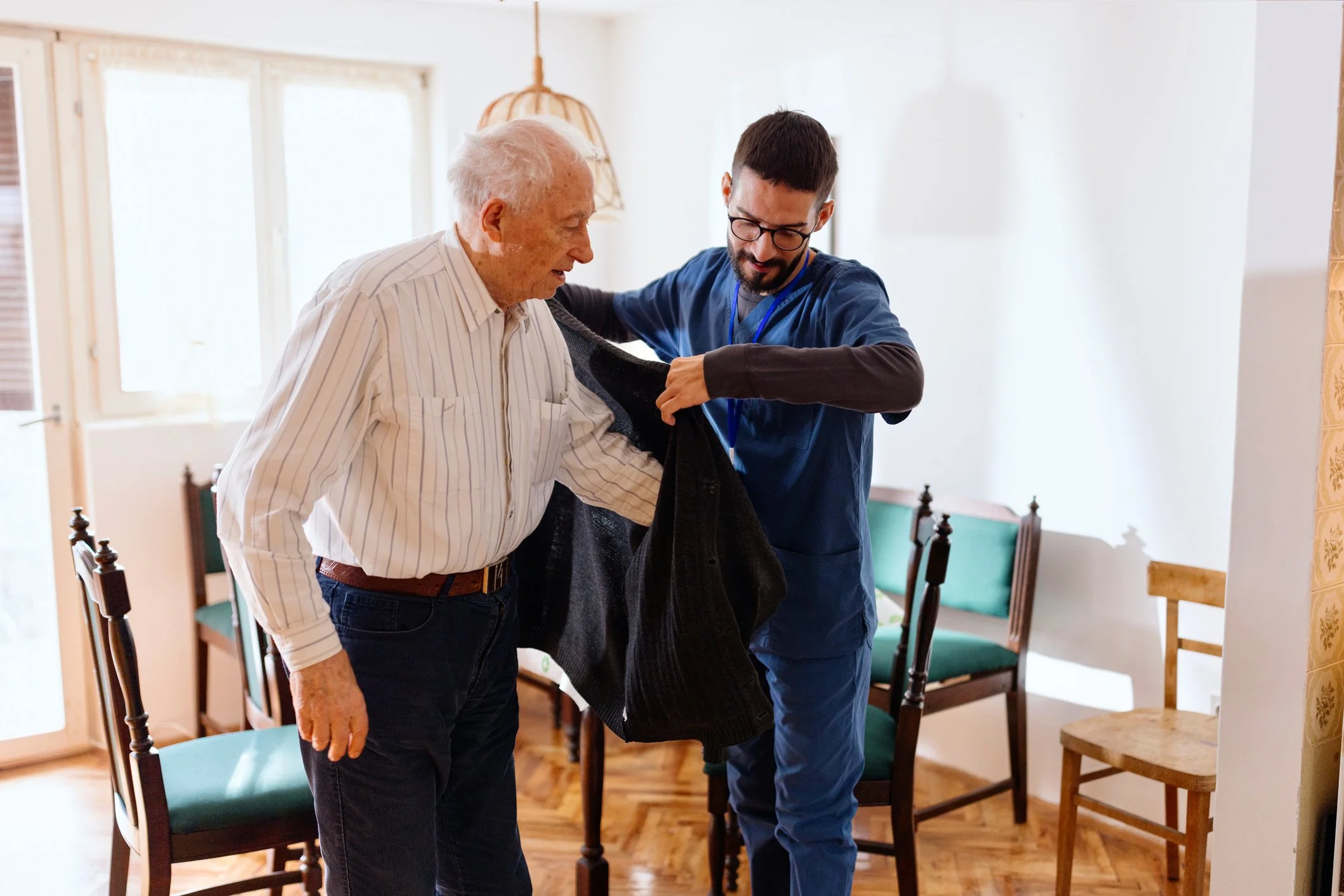 A caregiver helping an elderly man put on a black sweater in a bright room with wooden furniture.