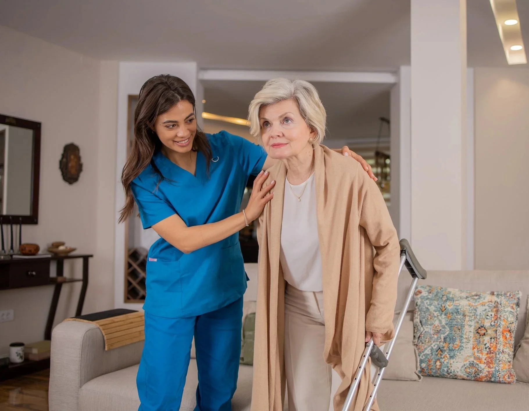 A young nurse in blue scrubs smiling and helping an elderly woman with a walker, who is wearing a beige coat, in a living room.