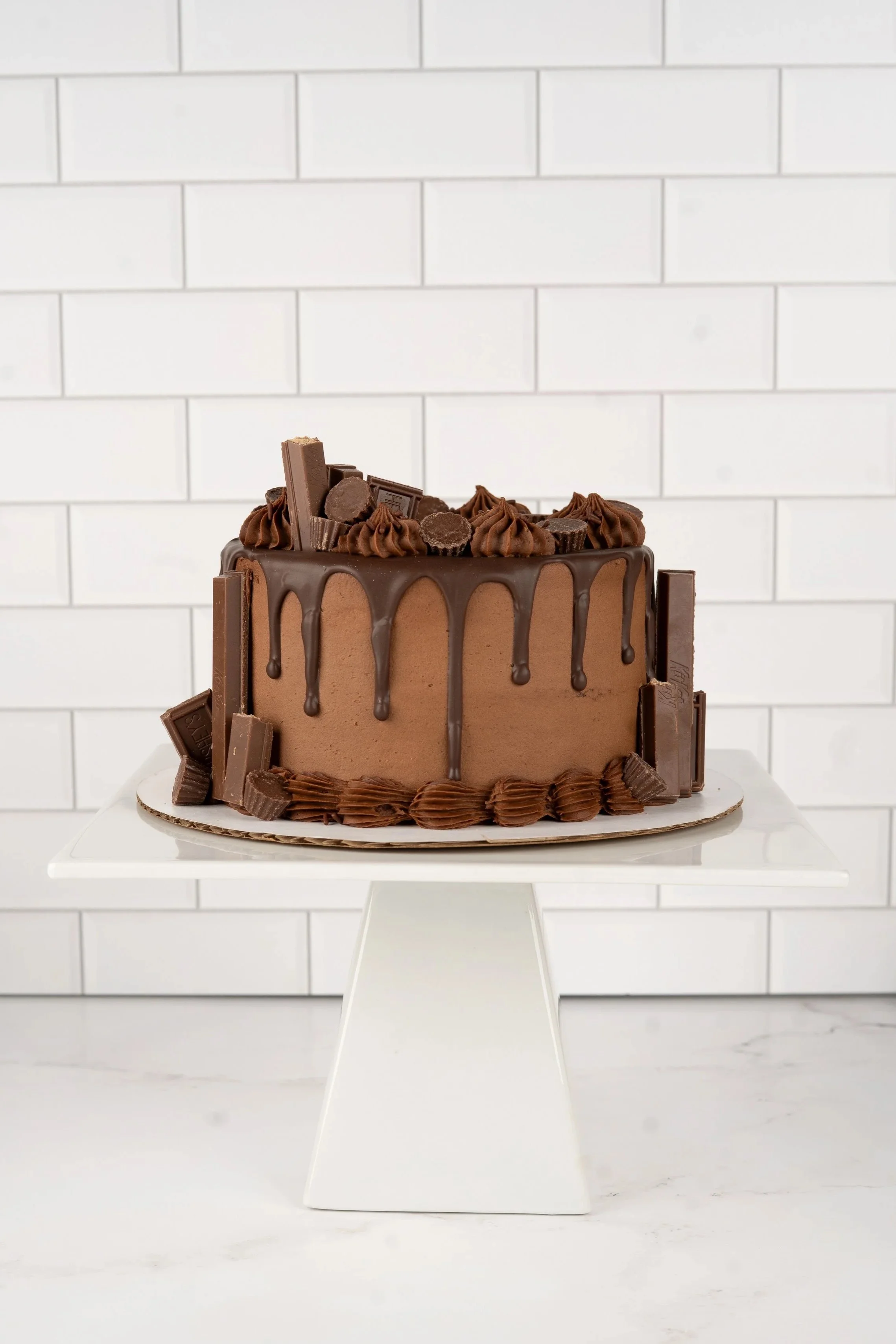 Chocolate cake with chocolate ganache drip, topped with chocolate pieces and piped chocolate frosting on white cake stand against a white tiled background.