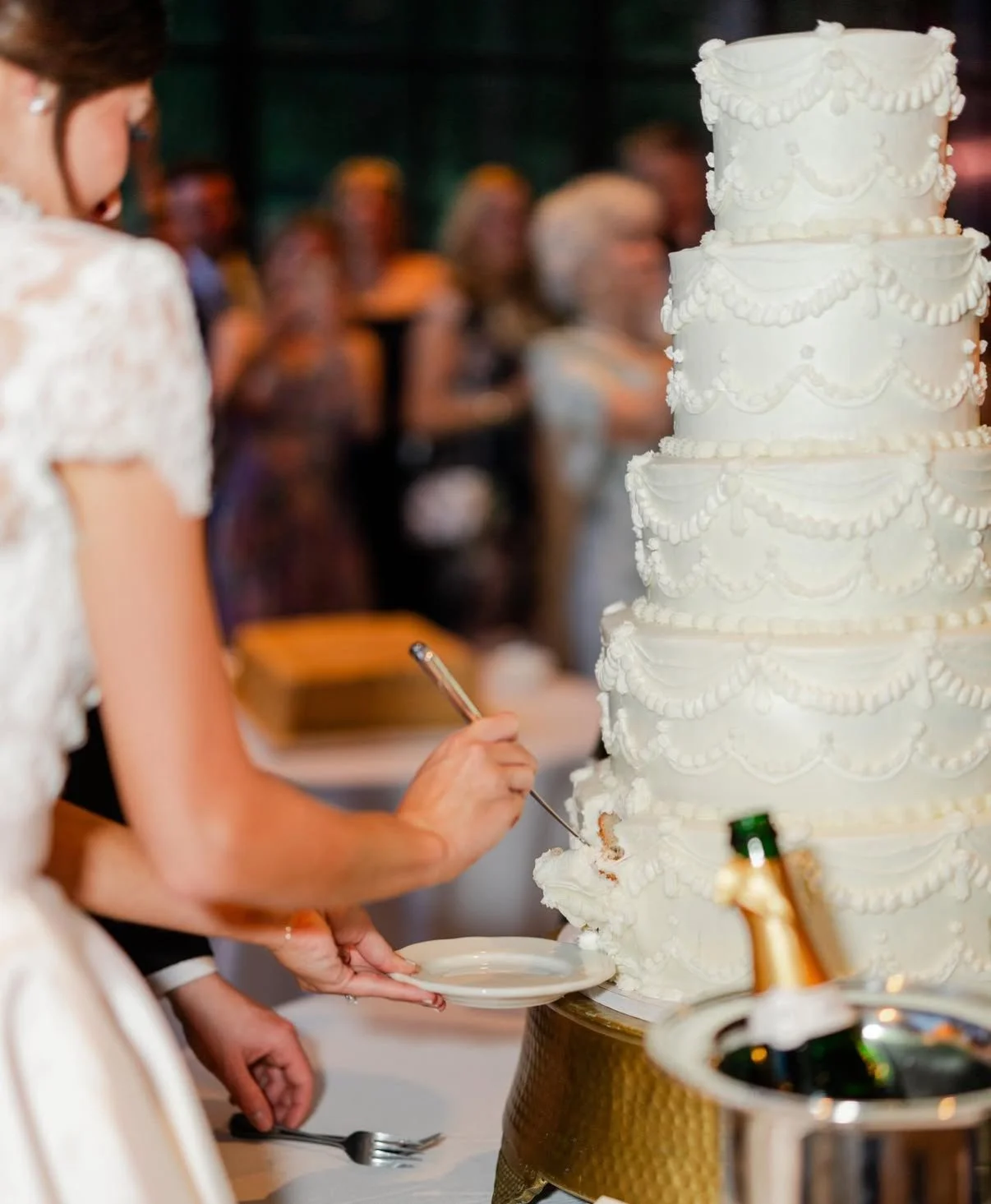 A woman in a white dress cuts a wedding cake on a table at a celebration, with guests in the background.