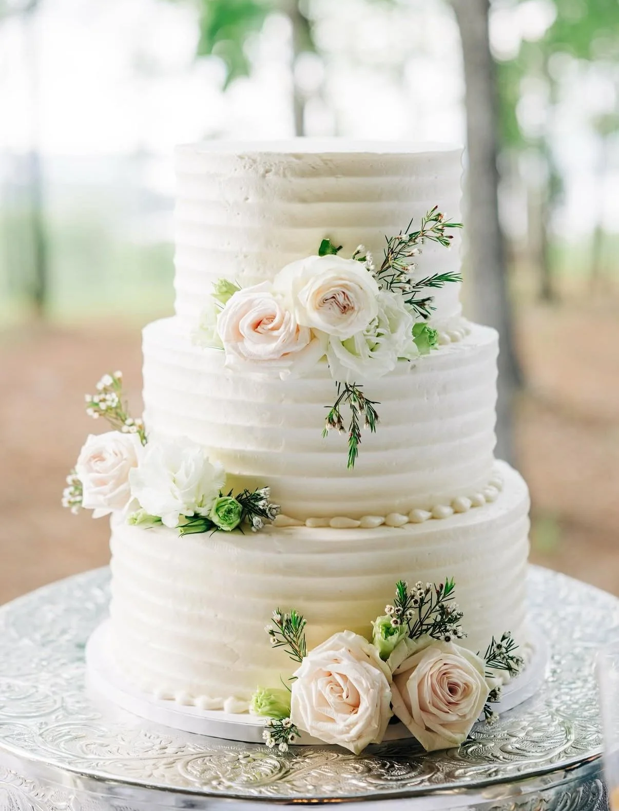 Three-tier white wedding cake decorated with white roses and greenery, set on a silver cake stand in an outdoor setting with trees in the background.