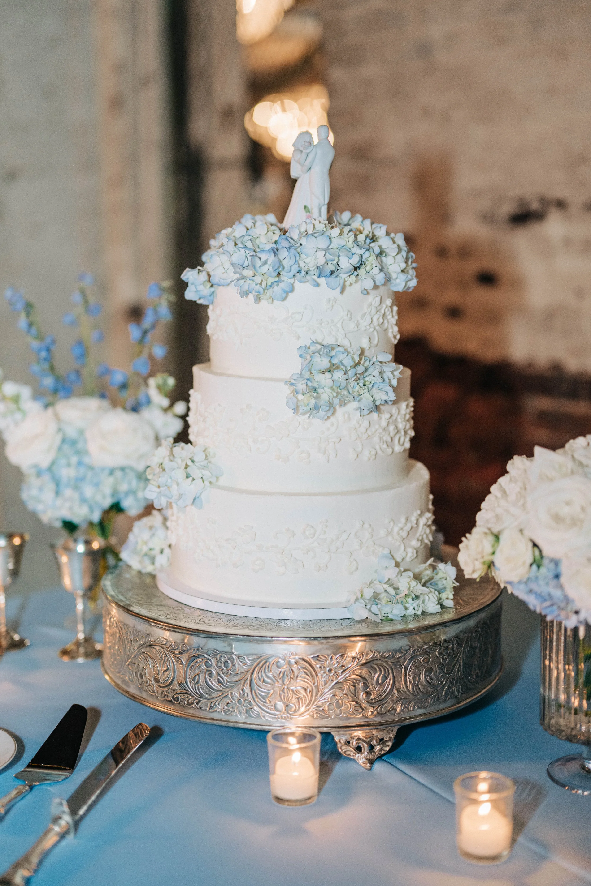 Three-tier white wedding cake decorated with white and light blue flowers, topped with a cake topper of a bride and groom, on an ornate silver cake stand, surrounded by candles and floral arrangements.