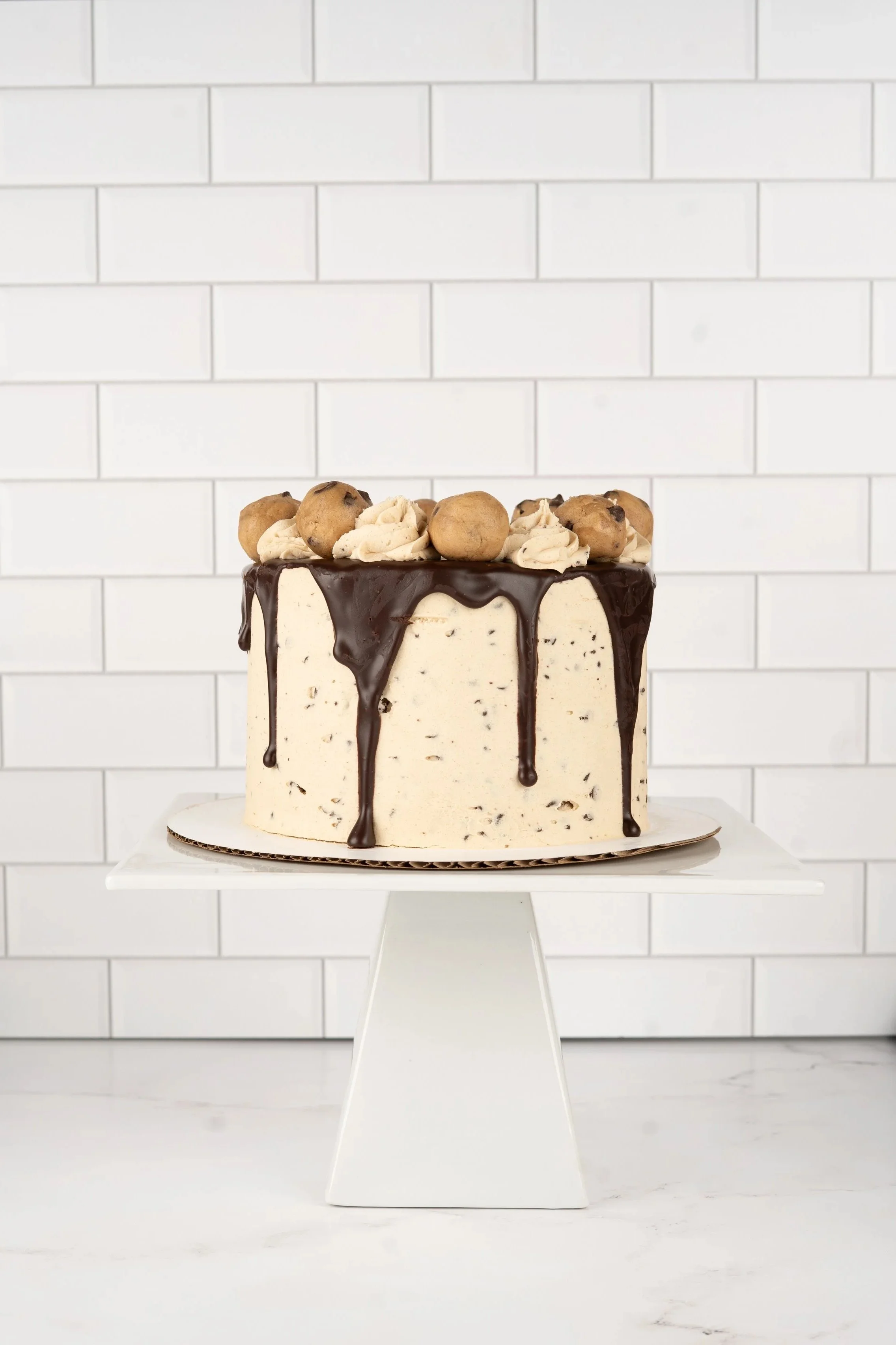 A cake with white frosting and chocolate drizzle on top, decorated with small cream puffs and cookies, placed on a white cake stand against a white tiled wall.