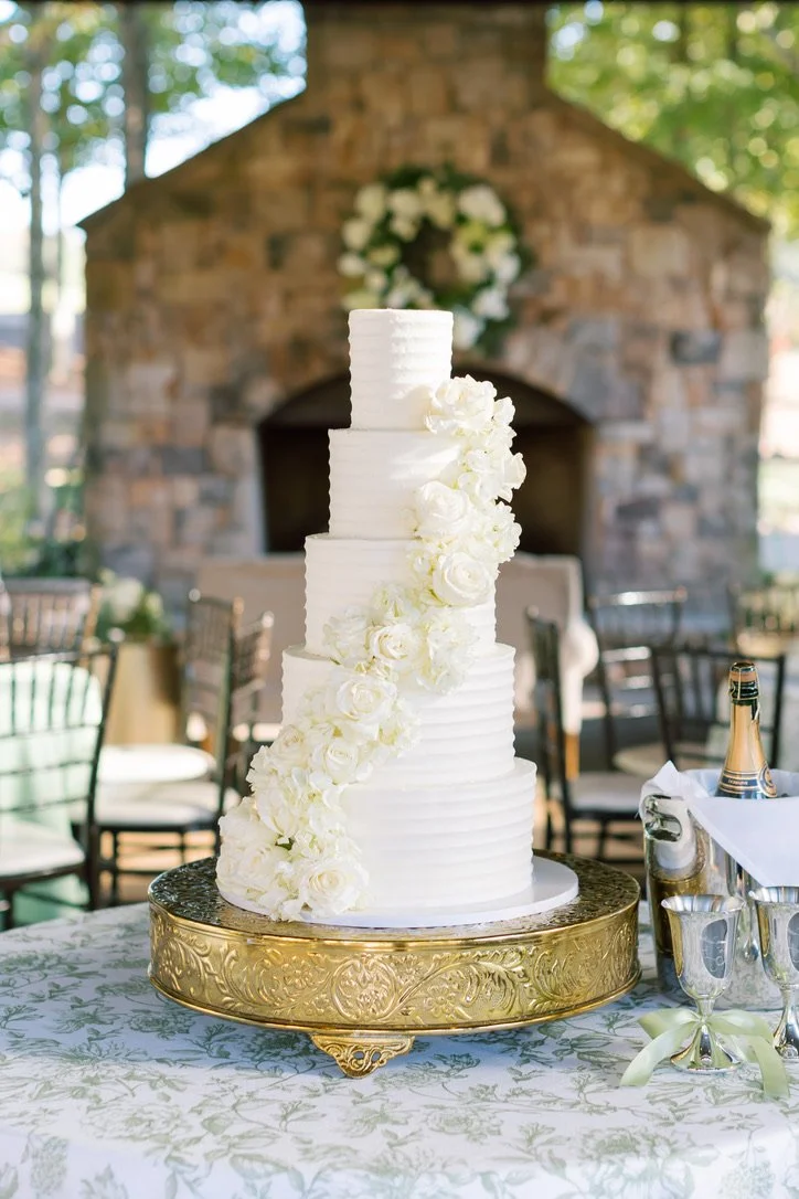 A tall, multi-tiered white wedding cake decorated with cascading white roses, placed on a gold ornate cake stand on a decorated table at an outdoor celebration.