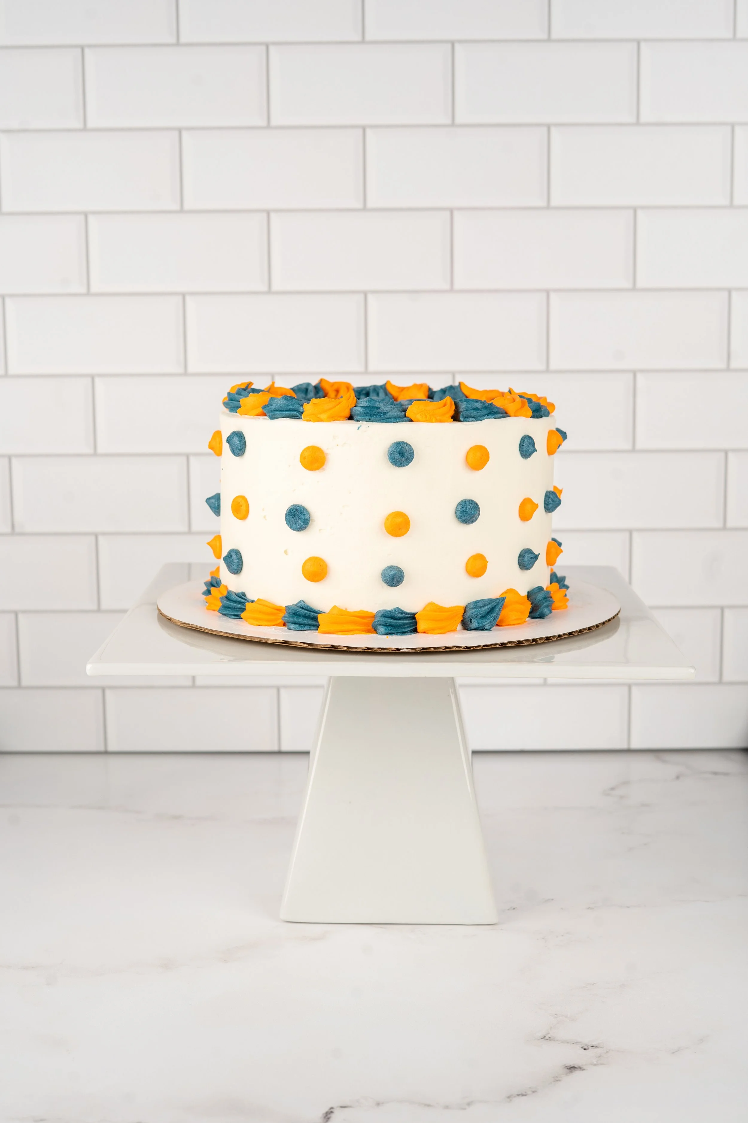 A white cake decorated with blue and orange buttercream dots and piping on a white cake stand.