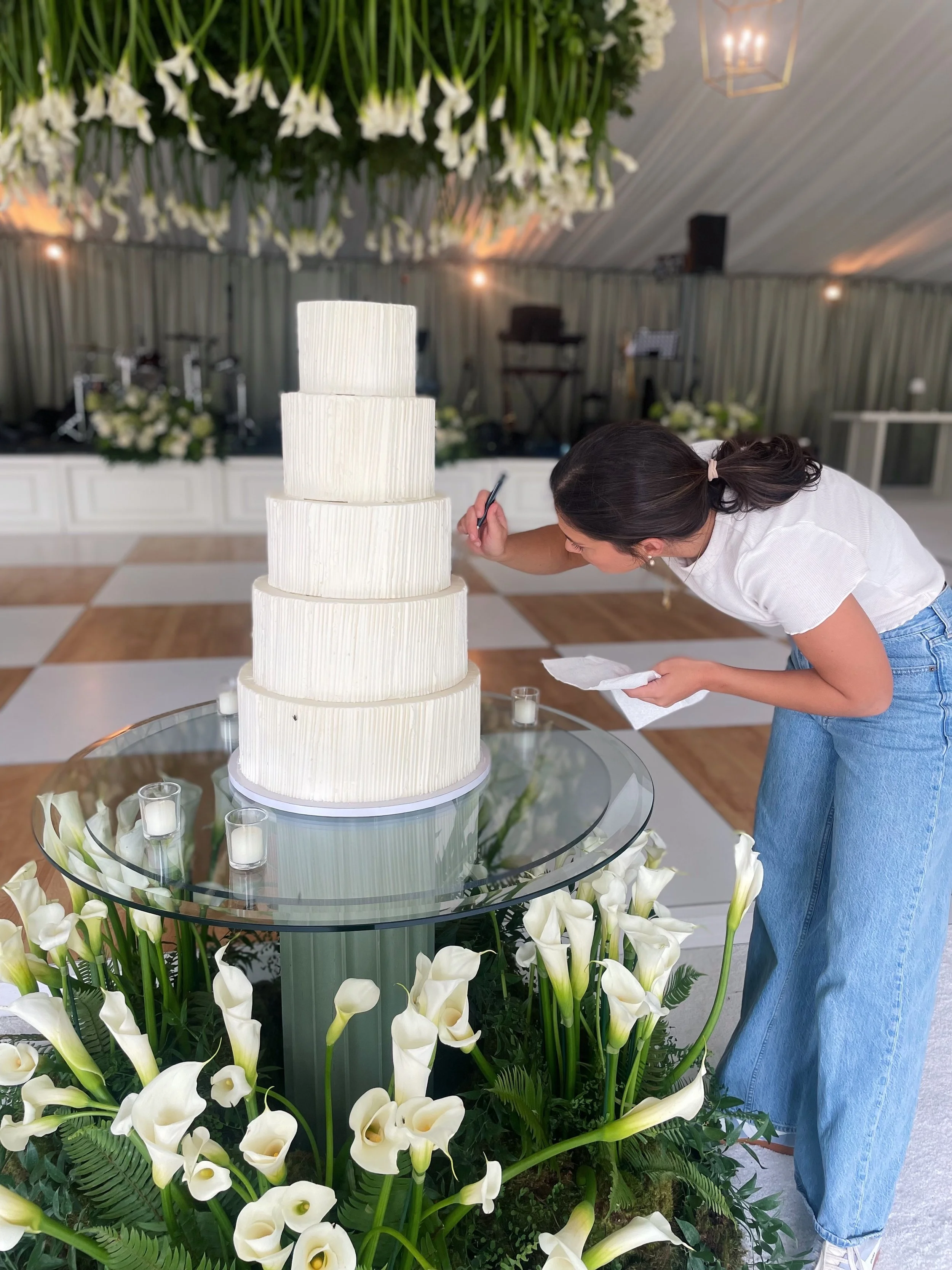 Woman decorating a multi-tiered white wedding cake at a wedding reception, surrounded by white calla lilies and candles.