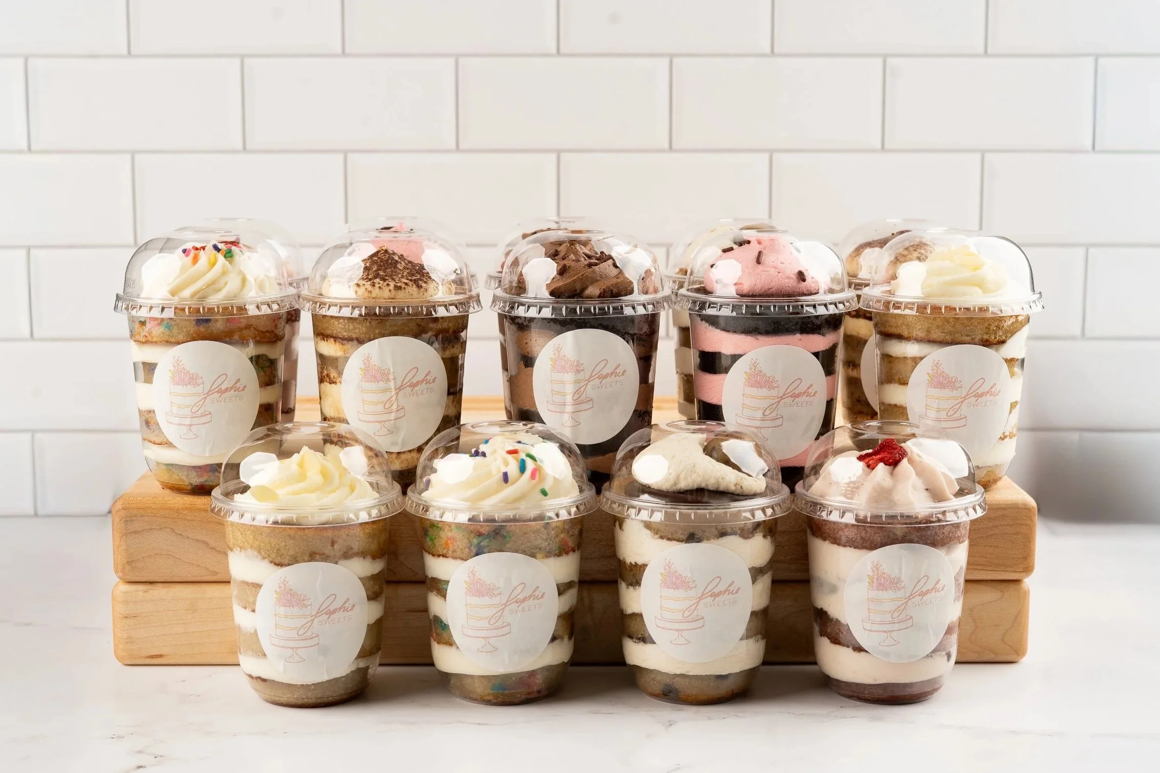 Assorted layered mini cheesecakes in clear plastic cups with clear domed lids, placed on a wooden surface with a white tiled background.