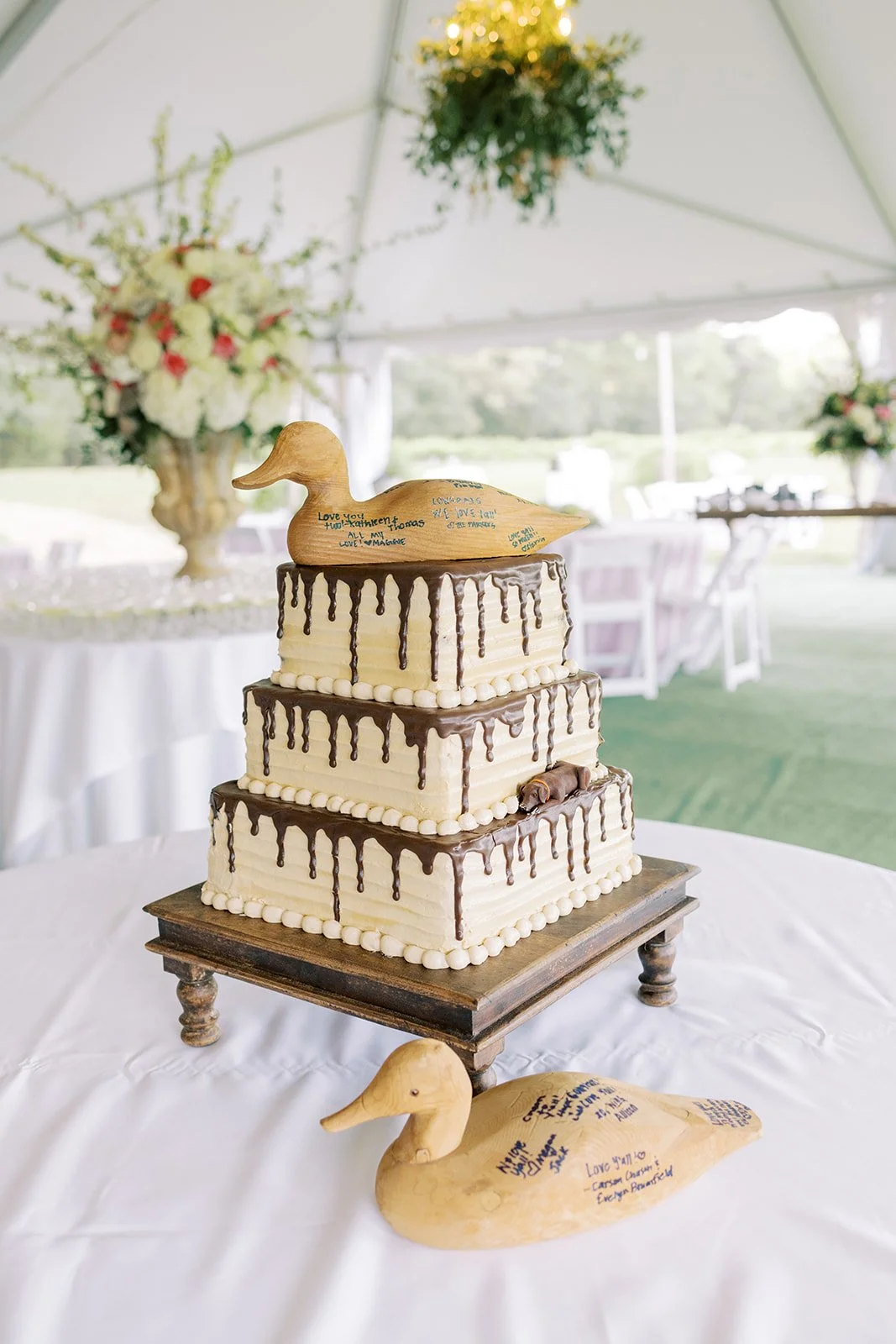 A three-tiered wedding cake decorated with chocolate drip and topped with a wooden duck figurine, with an additional duck figurine on the table in front, all decorated with handwritten messages, set in a wedding reception tent with floral arrangement