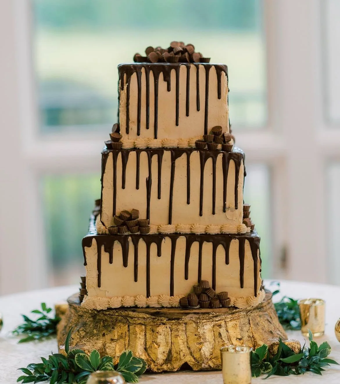 Three-tiered wedding cake with chocolate drip decoration, placed on a wooden cake stand, surrounded by greenery and gold candleholders.