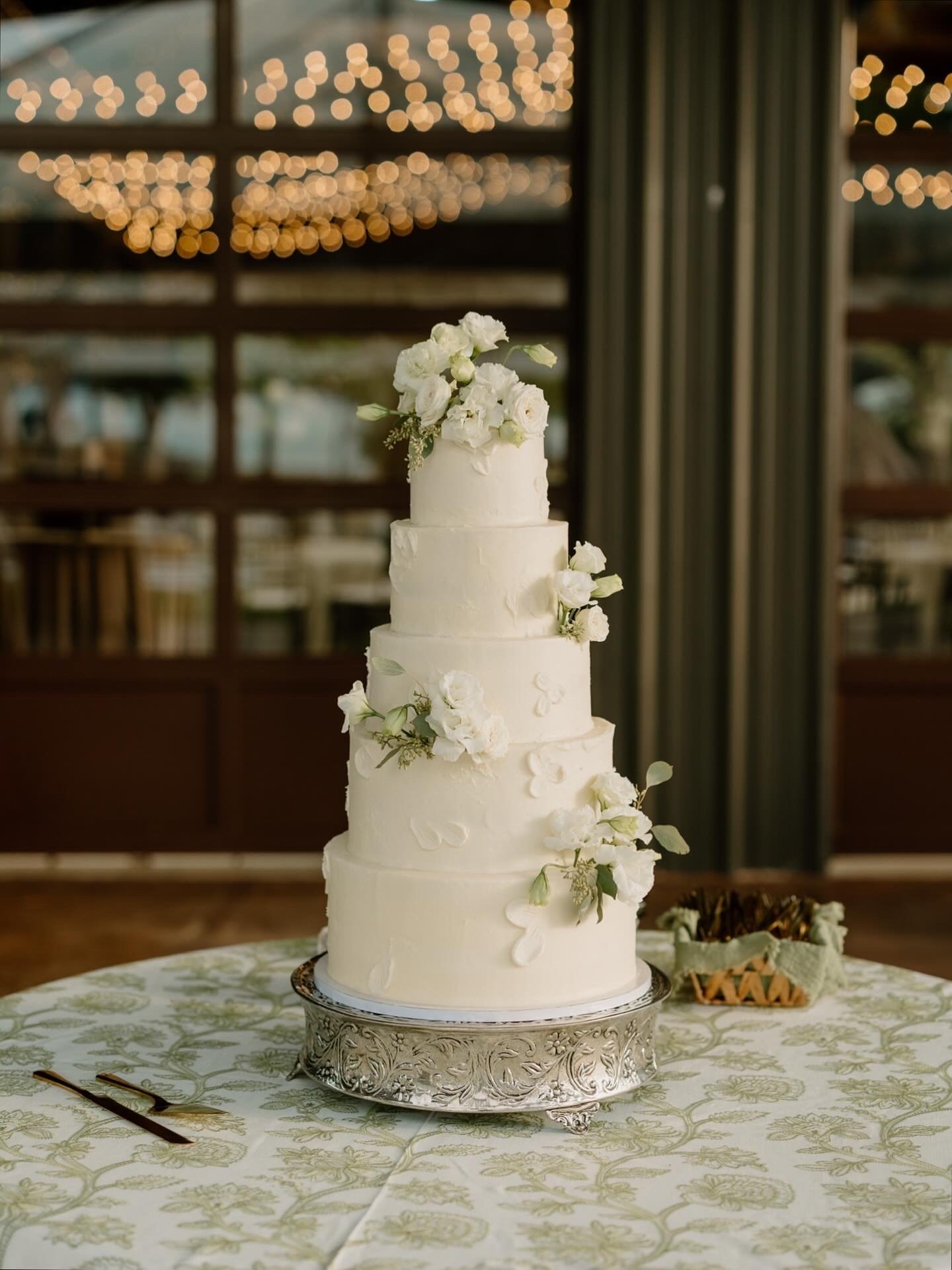 White five-tier wedding cake with floral decorations on a silver cake stand, set on a table with a patterned tablecloth. The background features warm string lights and dark curtains.