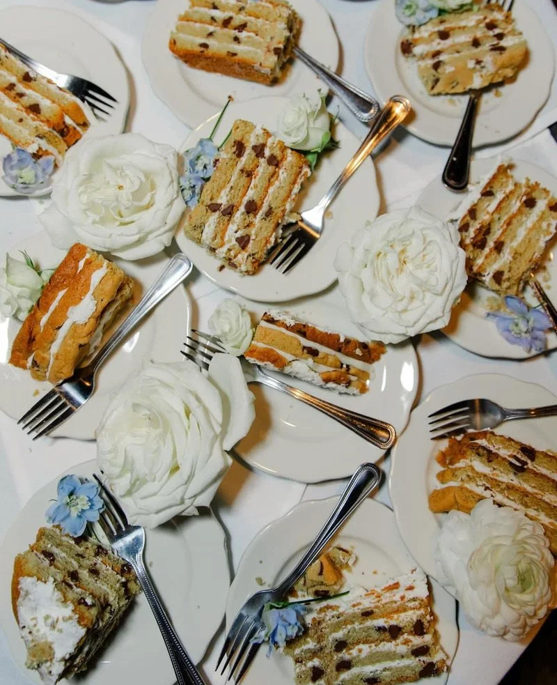 Multiple plates of layered cake slices with white frosting, some garnished with edible flowers, and dollops of whipped cream.