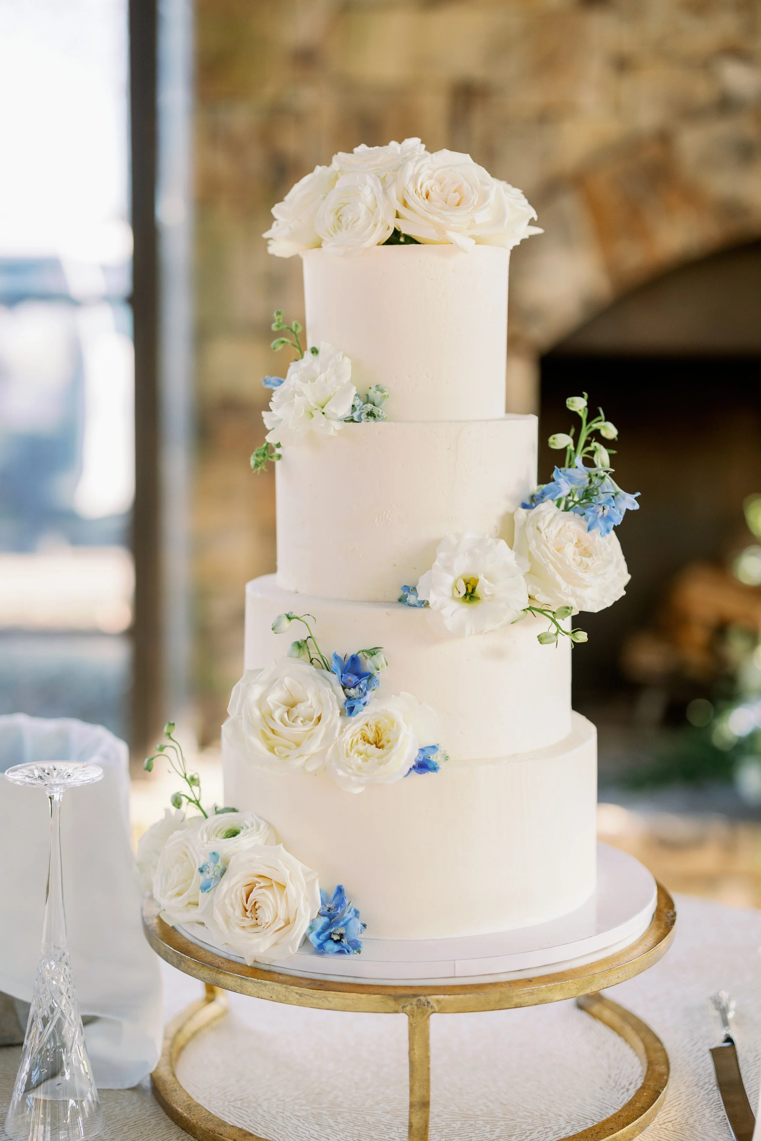 Four-tier white wedding cake decorated with white roses, white lisianthus, and blue flowers, placed on a gold stand.