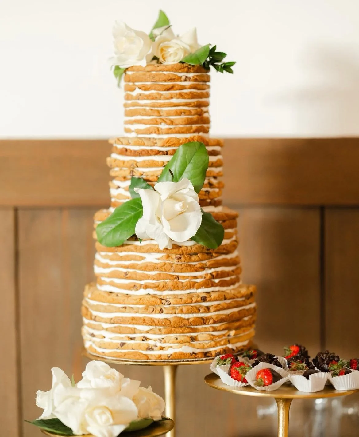 A tall, tiered cookie cake decorated with white flowers and green leaves, set on a gold stand, surrounded by strawberries and blackberries in white paper cups.