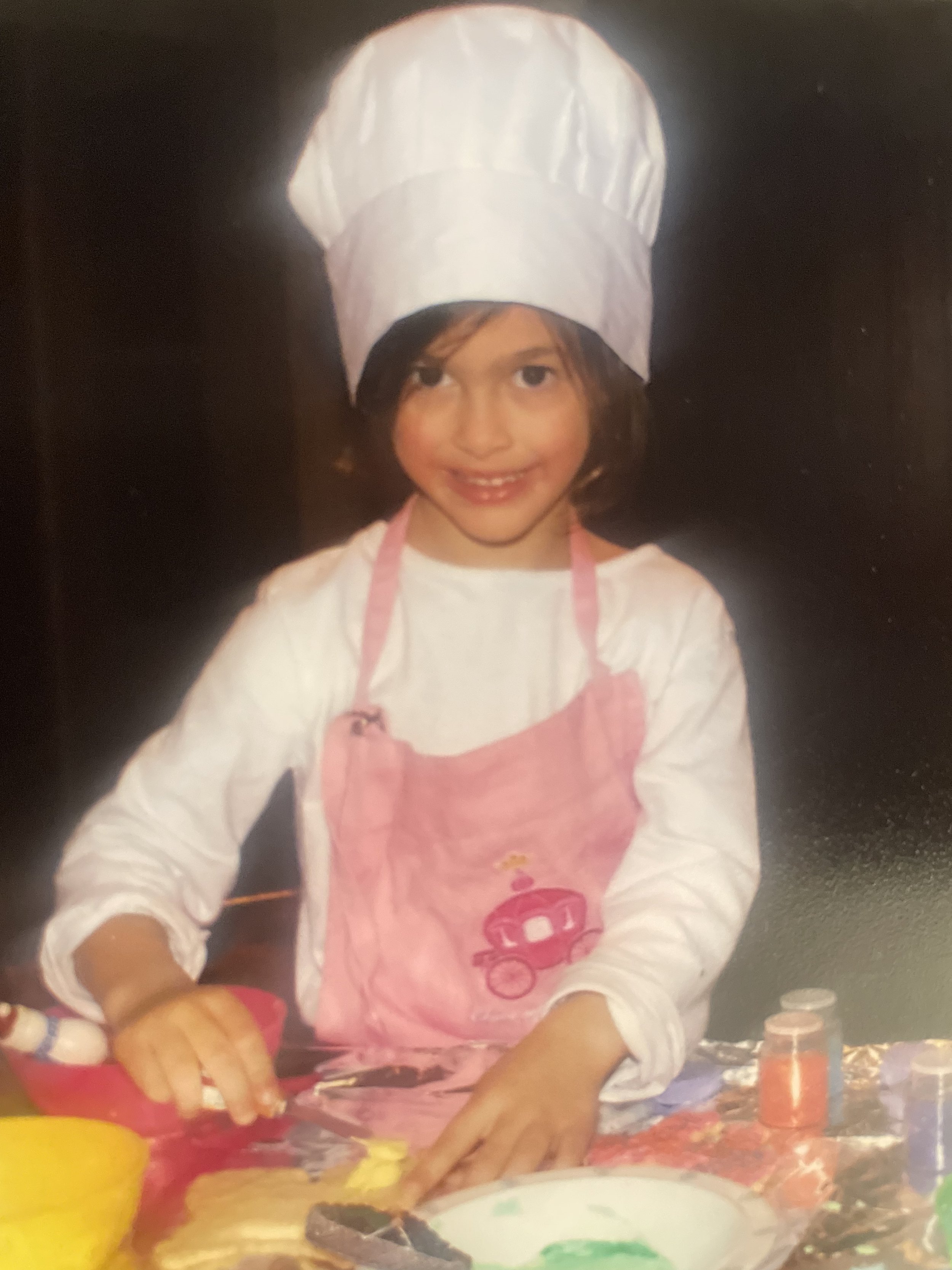 A young girl dressed as a chef, wearing a white chef hat and pink apron, cooking or baking with colorful ingredients on a table.