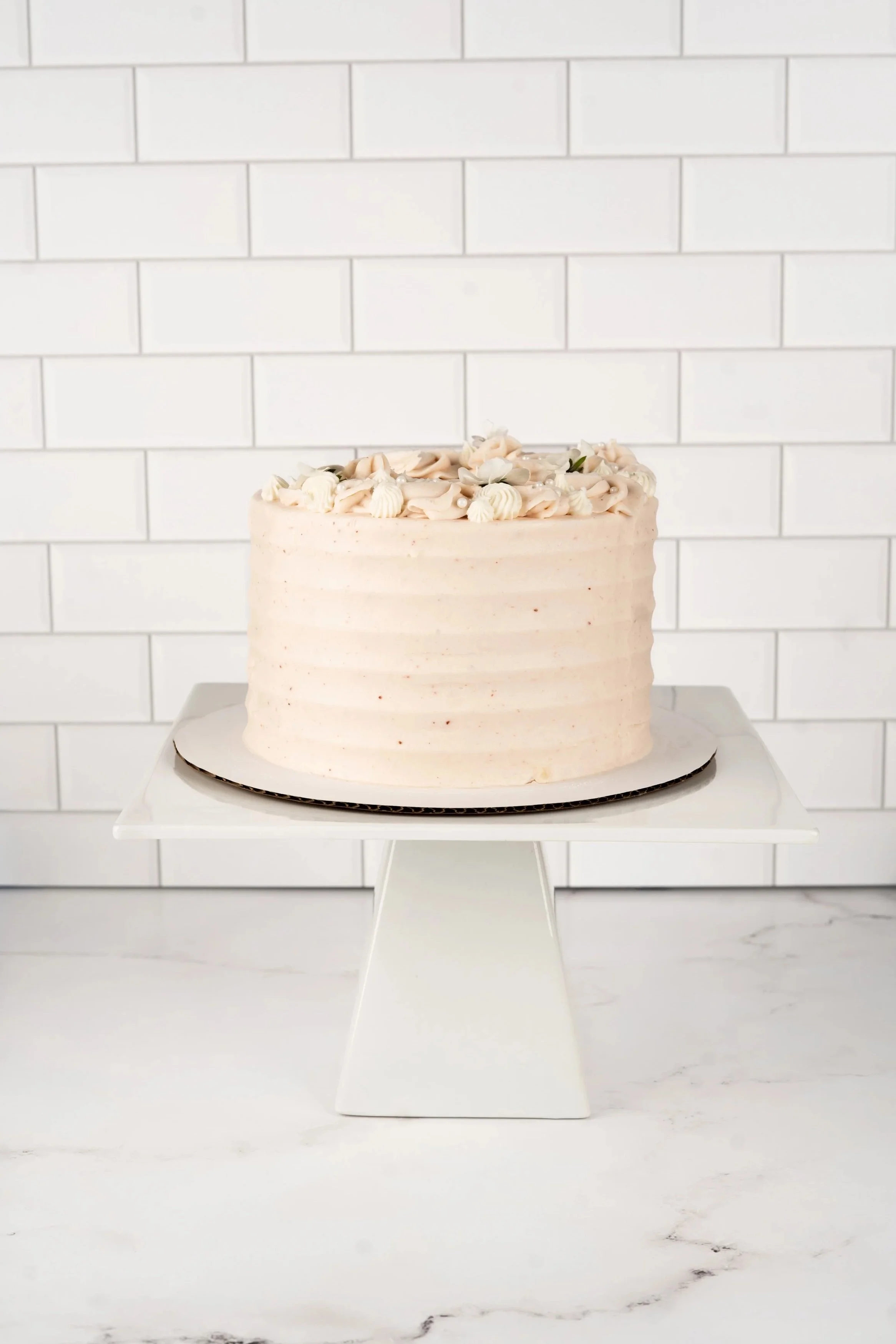 A white frosted layered cake decorated with swirl and shell piping on top, placed on a white cake stand against a white tile background.