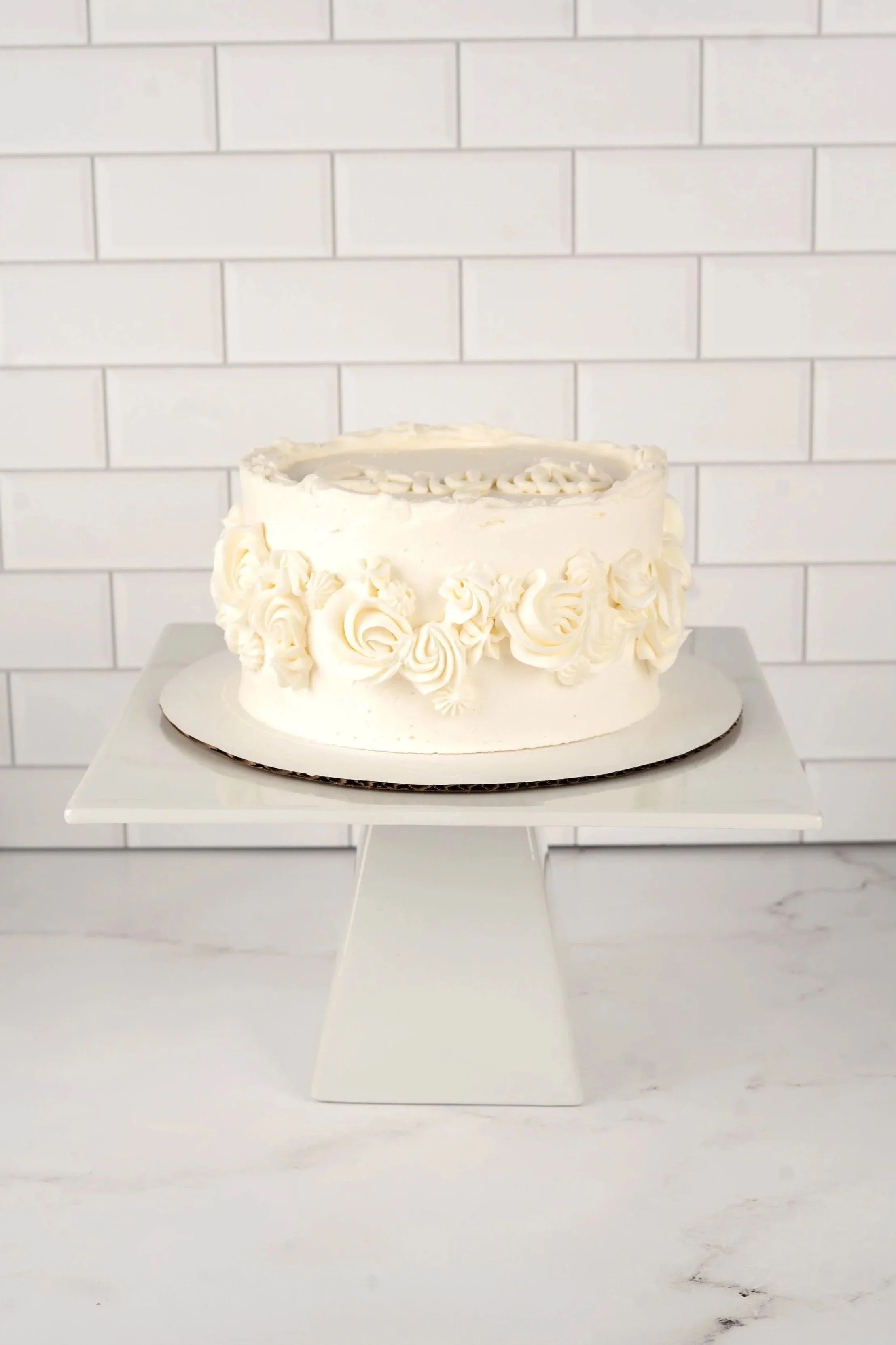 A round white cake with decorative floral piping, placed on a white cake stand in front of a white brick wall.