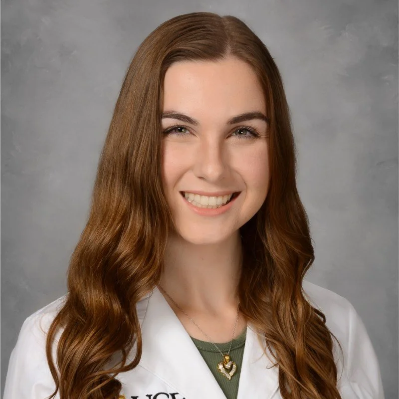 Professional portrait of a young, female physical therapist with long, wavy auburn hair, smiling, wearing a white lab coat and a necklace with a heart-shaped pendant.