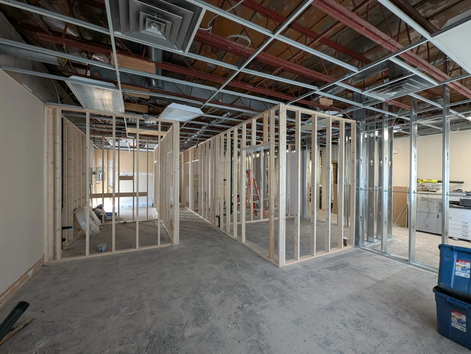 Interior of a building under construction with wooden framing for walls and exposed ceiling infrastructure, including ductwork and lighting fixtures.