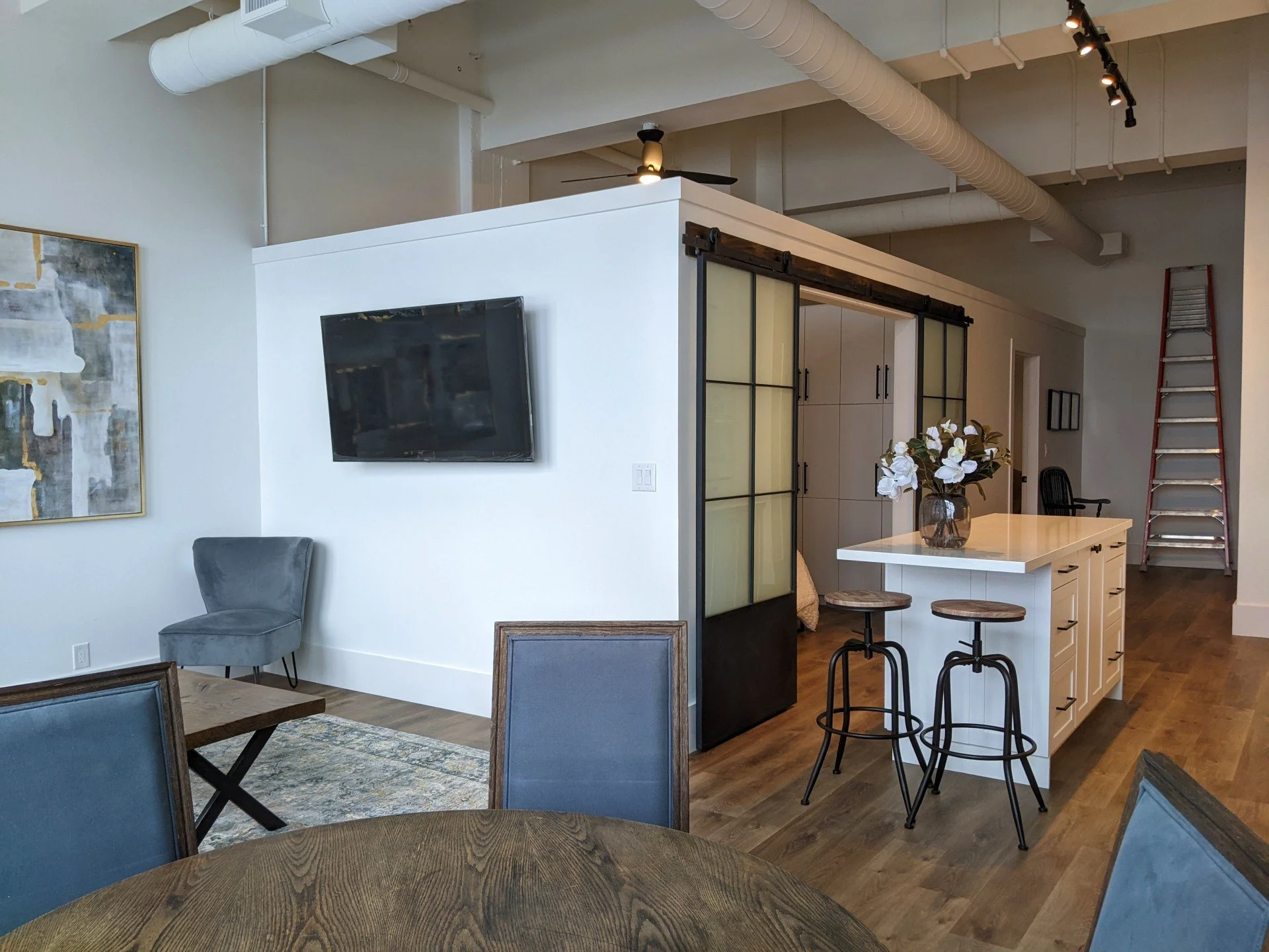 Modern living space with a circular dining table, blue upholstered chairs, a small gray armchair, a wall-mounted TV, a kitchen island with stools, a vase of white flowers, sliding barn doors, and a tall ladder in the background.