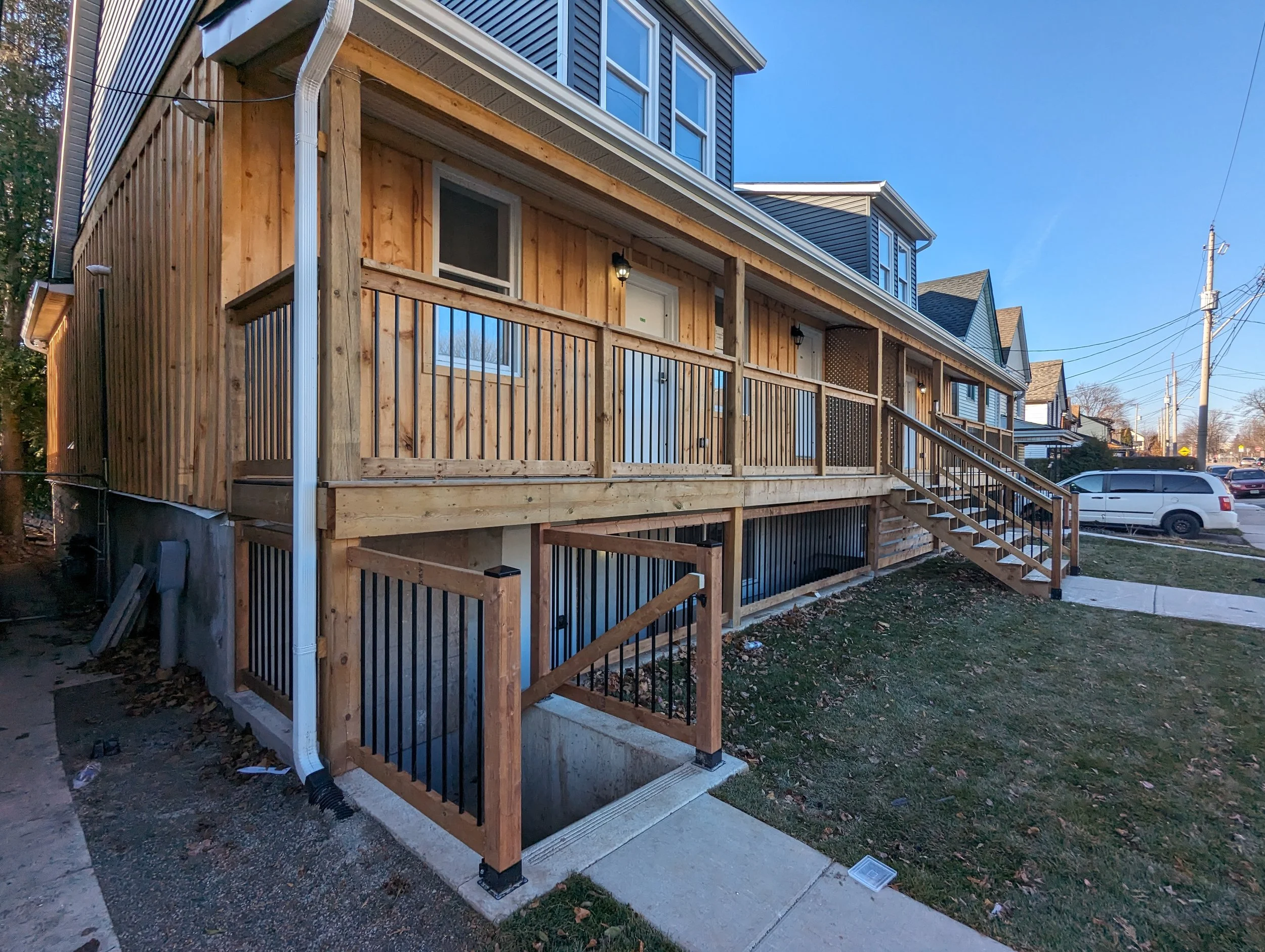 Front porch of a new wooden deck with stairs, metal railings, and some outdoor lighting, attached to a multi-story residential building, with a sidewalk and parked cars on the street.