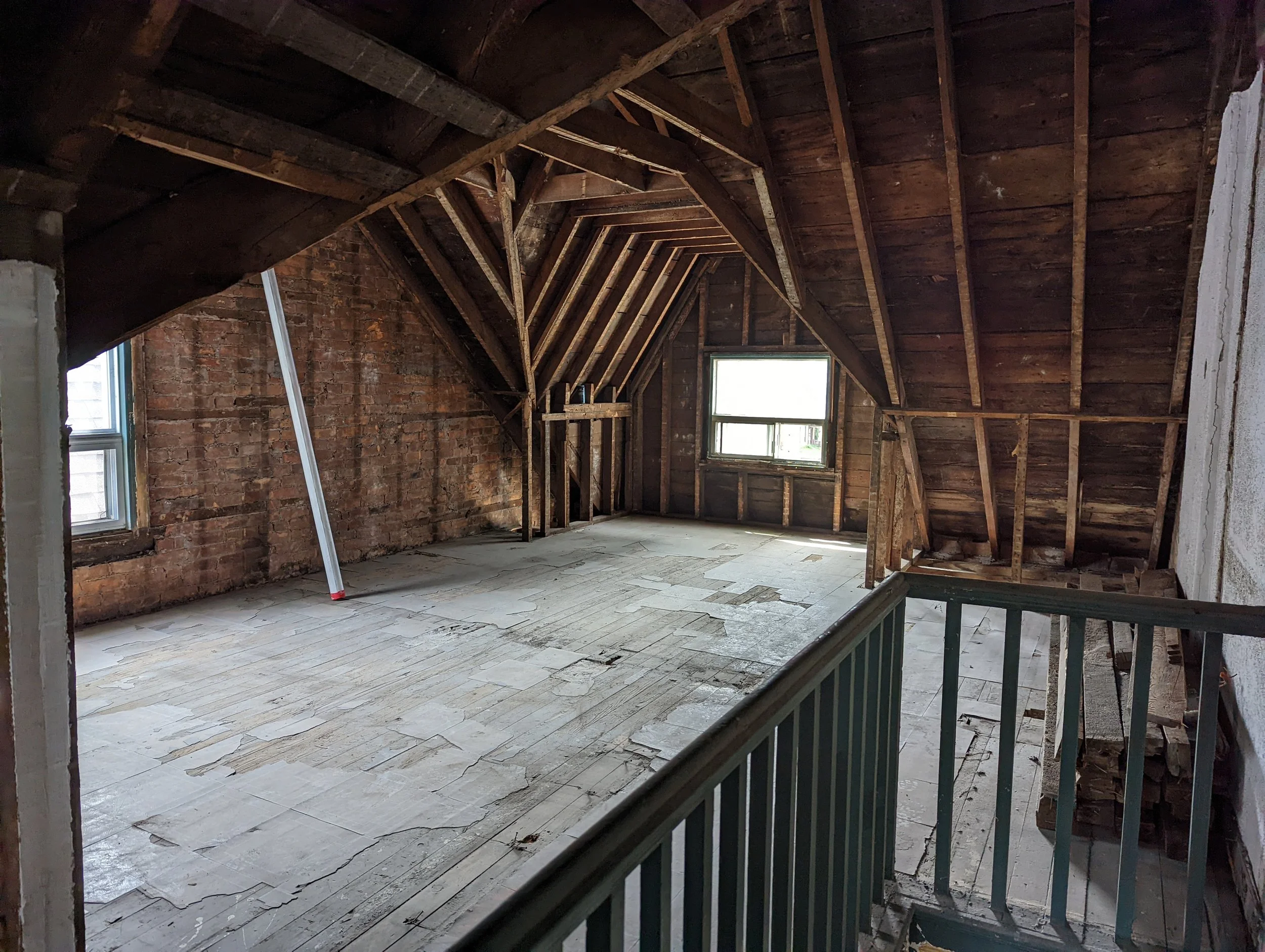 Empty attic space with exposed wooden beams, brick wall, two windows, and flooring under renovation.