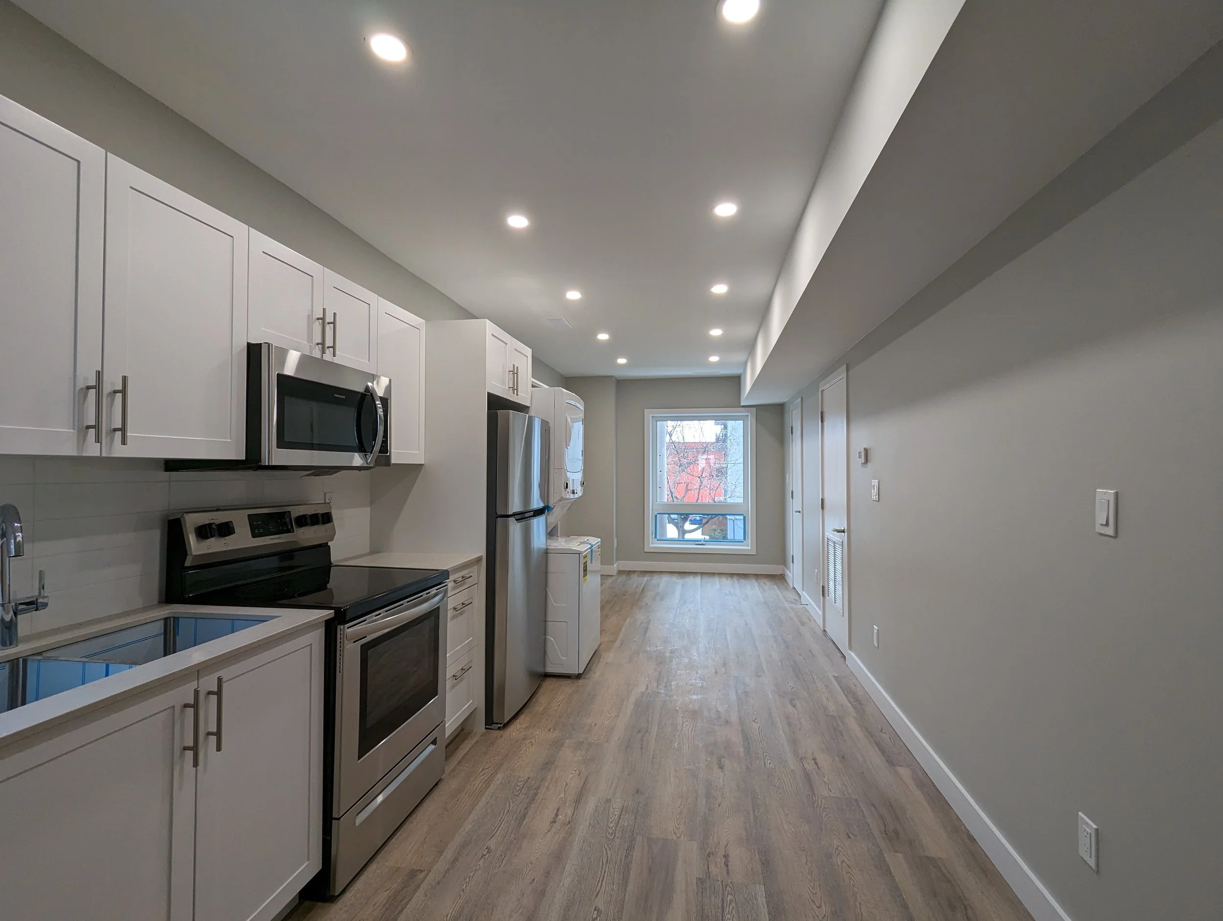 Modern kitchen with white cabinets, stainless steel appliances, wood flooring, and a window at the end.