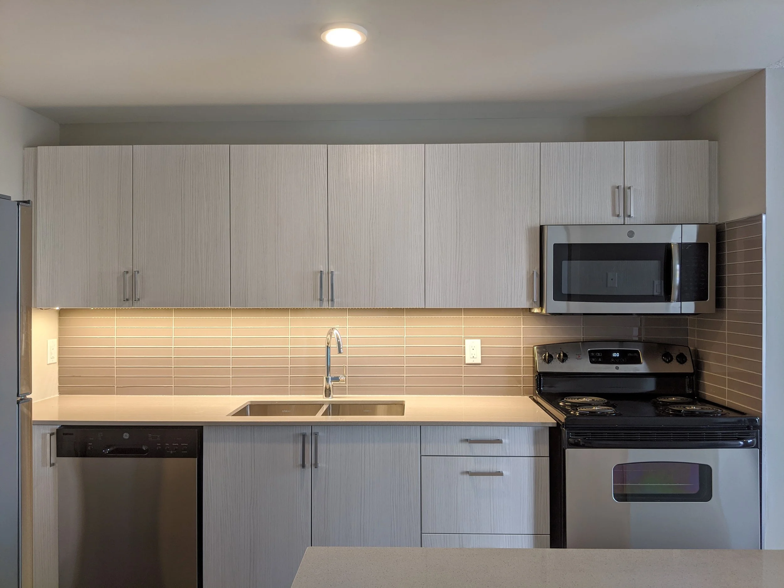 Modern kitchen with gray cabinets, beige countertops, a black stove, a stainless steel microwave, a dishwasher, and a tiled backsplash