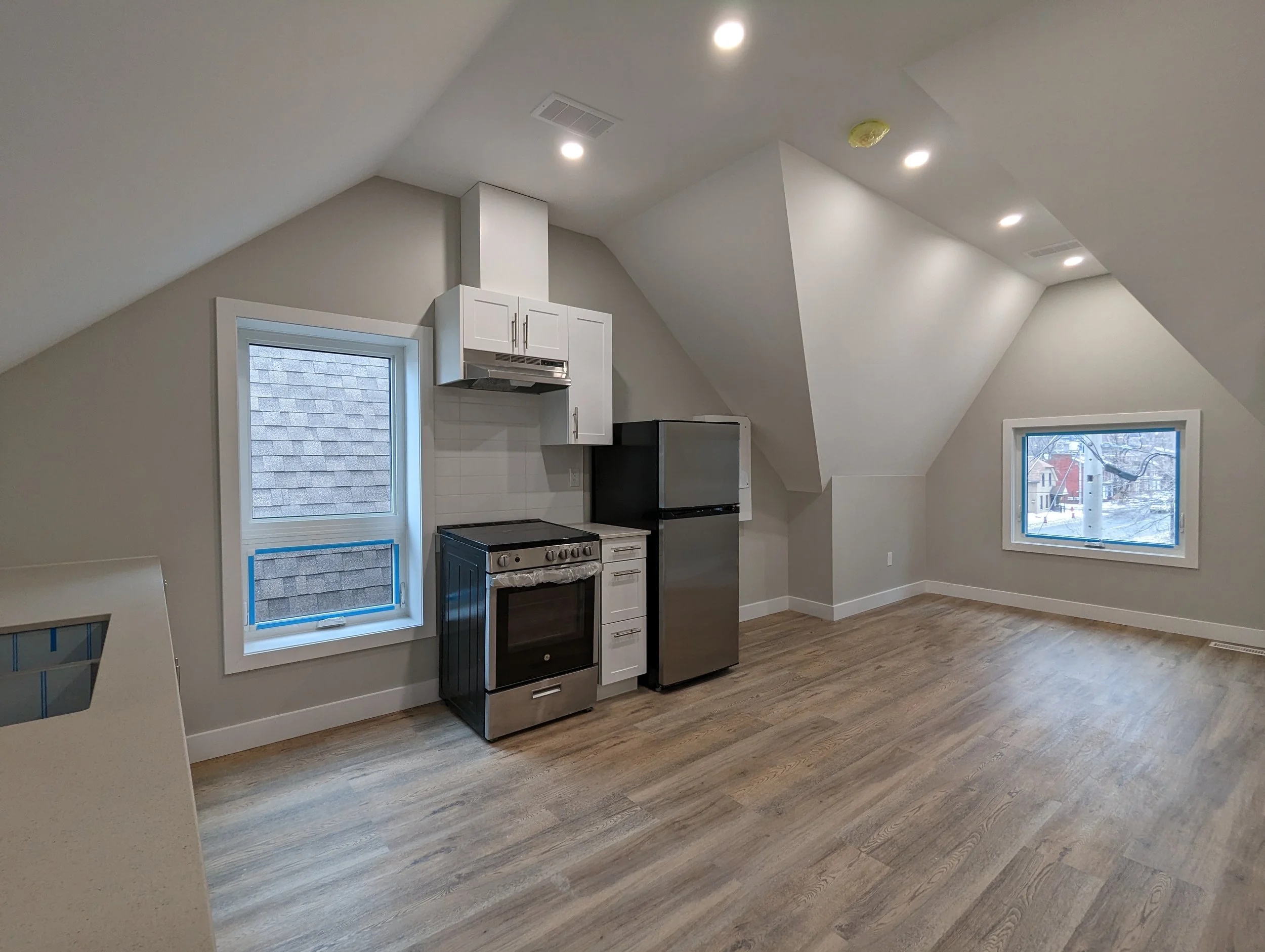 Empty kitchen with hardwood floors, white cabinets, a black stove, a stainless steel refrigerator, and two windows showing neighborhood outside.