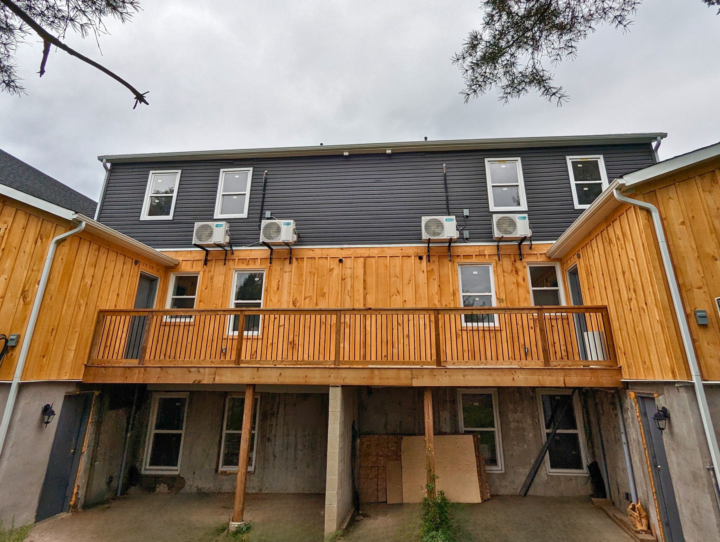 Construction site of a multi-story house with a wooden deck, black and wooden siding, multiple windows, and air conditioning units on the exterior wall.
