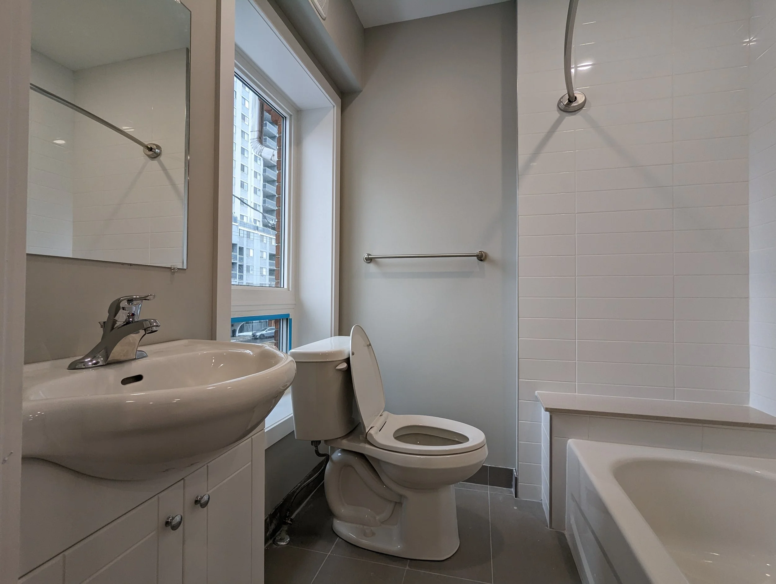 Bathroom with a white toilet, sink, mirror, and bathtub. A window shows a cityscape of tall buildings.