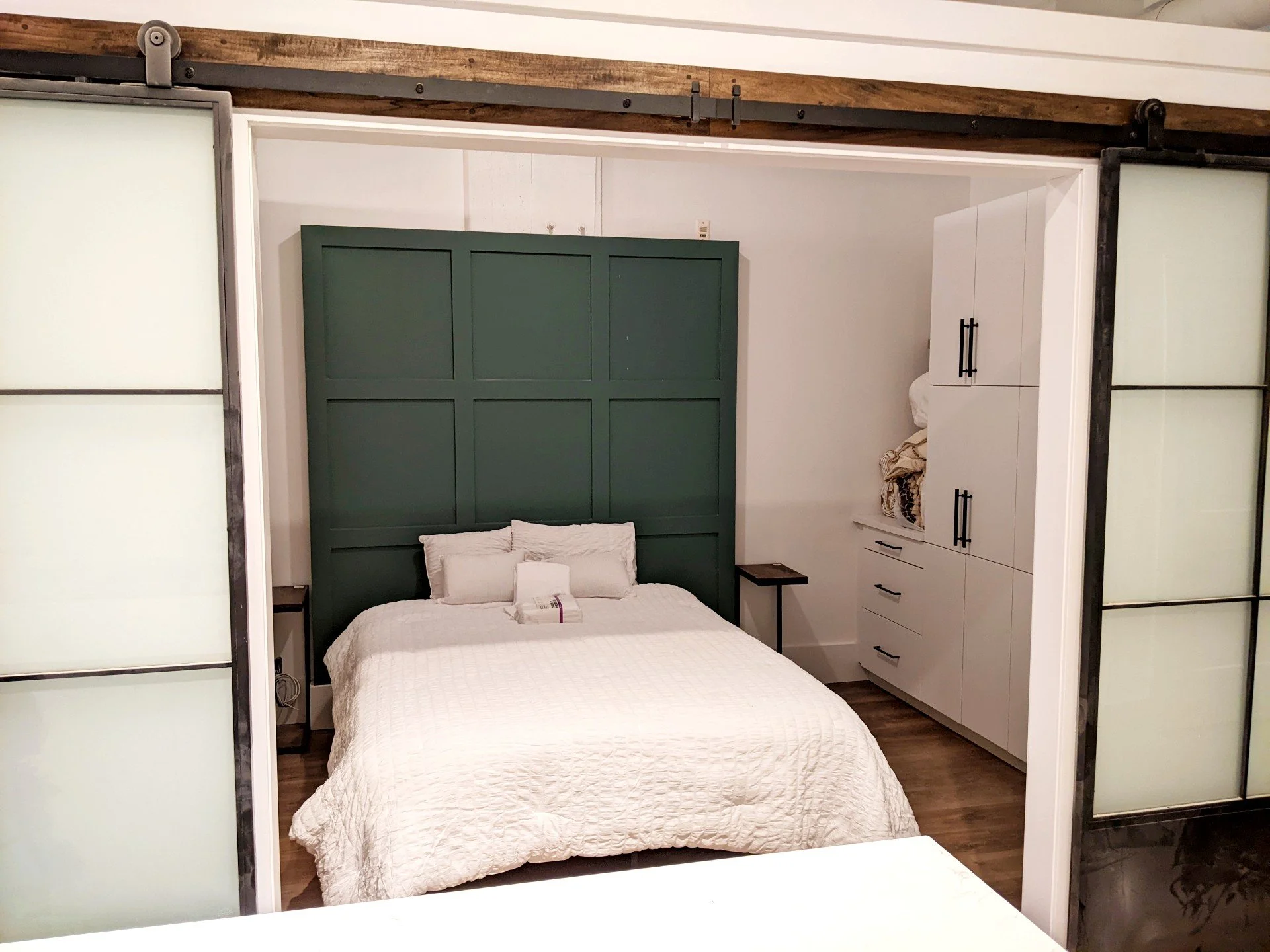 A bedroom with a bed featuring a dark green headboard, white bedspread, and pillows. To the right, there is a white built-in cabinet. The room has sliding doors with frosted glass panels.
