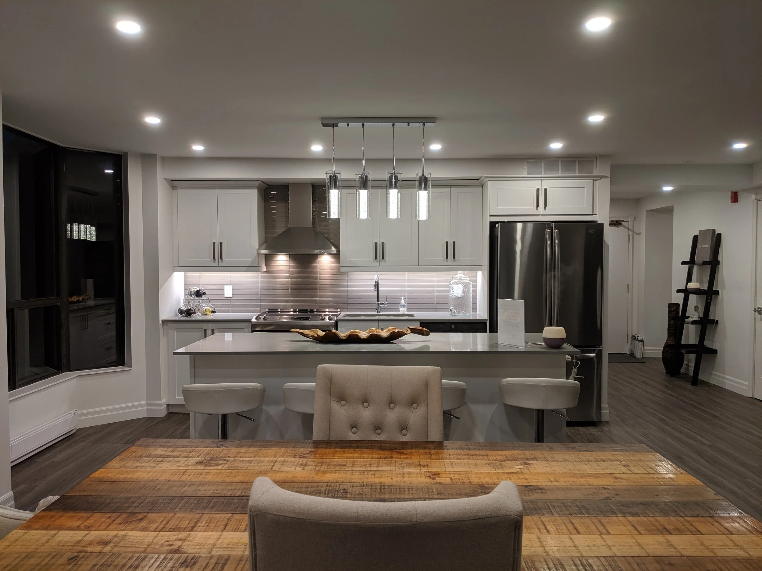 Modern kitchen with gray cabinets, stainless steel appliances, a large white island with bar stools, and a wooden dining table in the foreground.