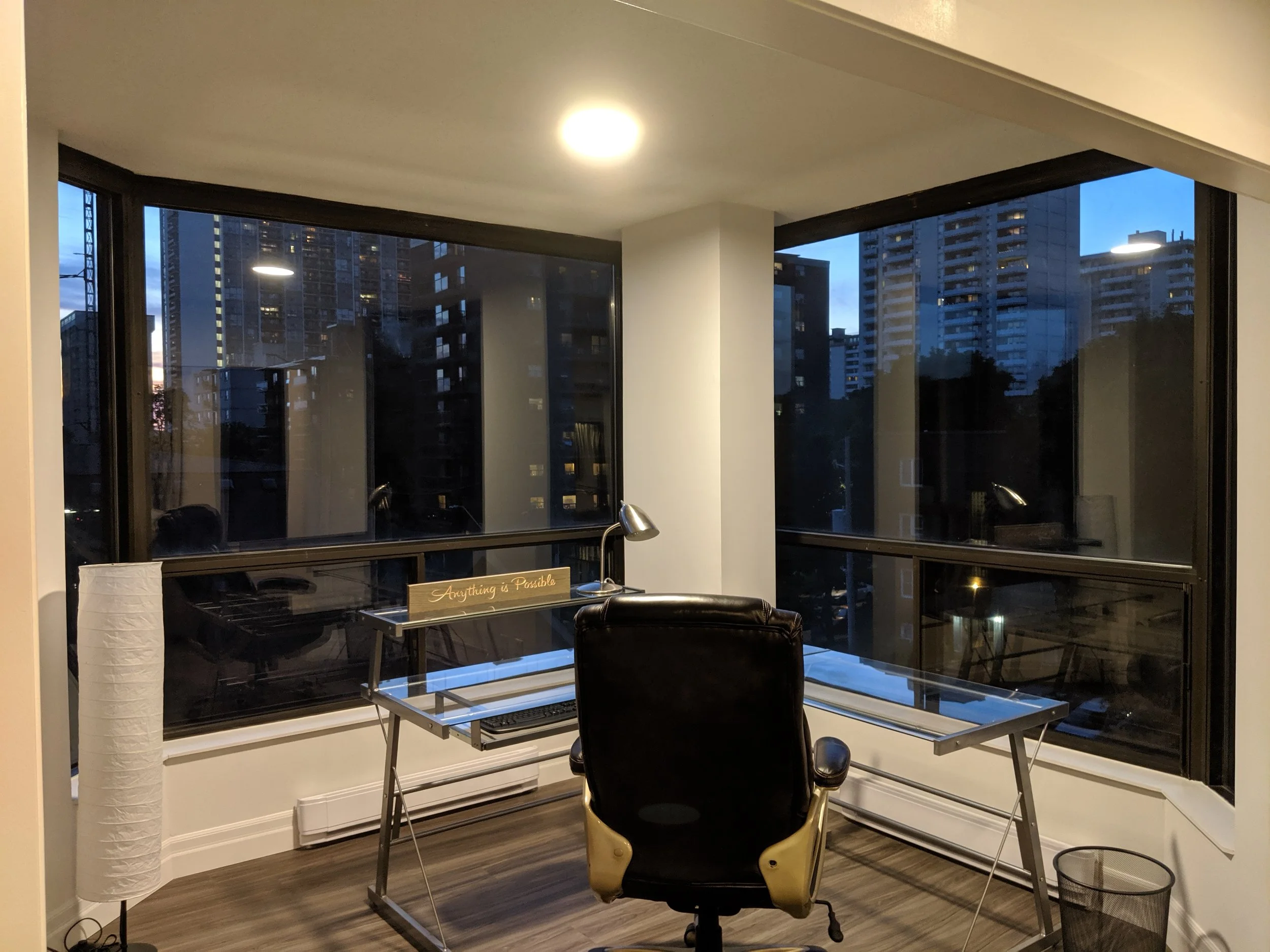Modern home office with a glass desk, black leather chair, and large corner windows showing city buildings at dusk.