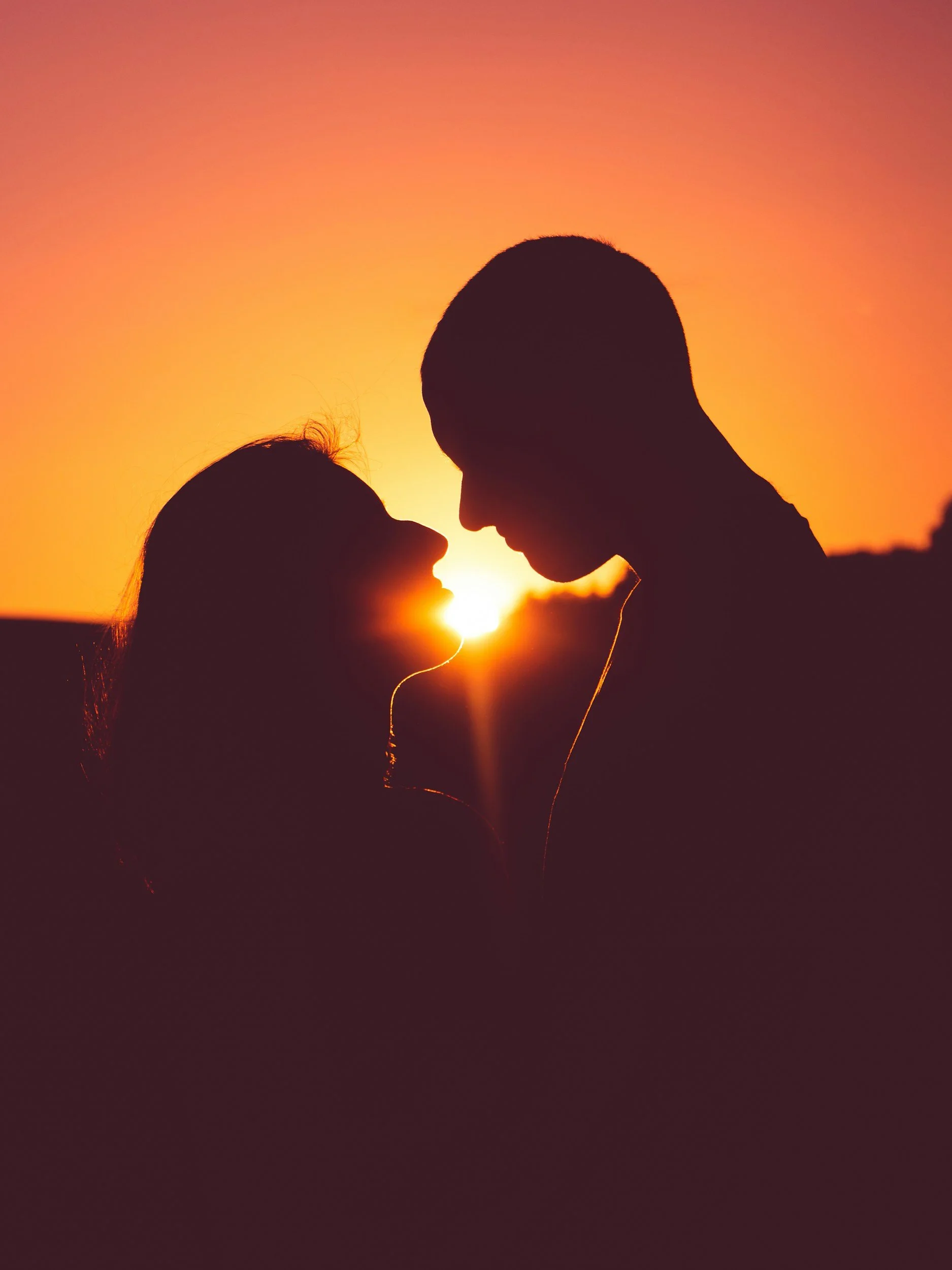 Silhouettes of a man and woman with their foreheads touching, set against a vibrant sunset sky.
