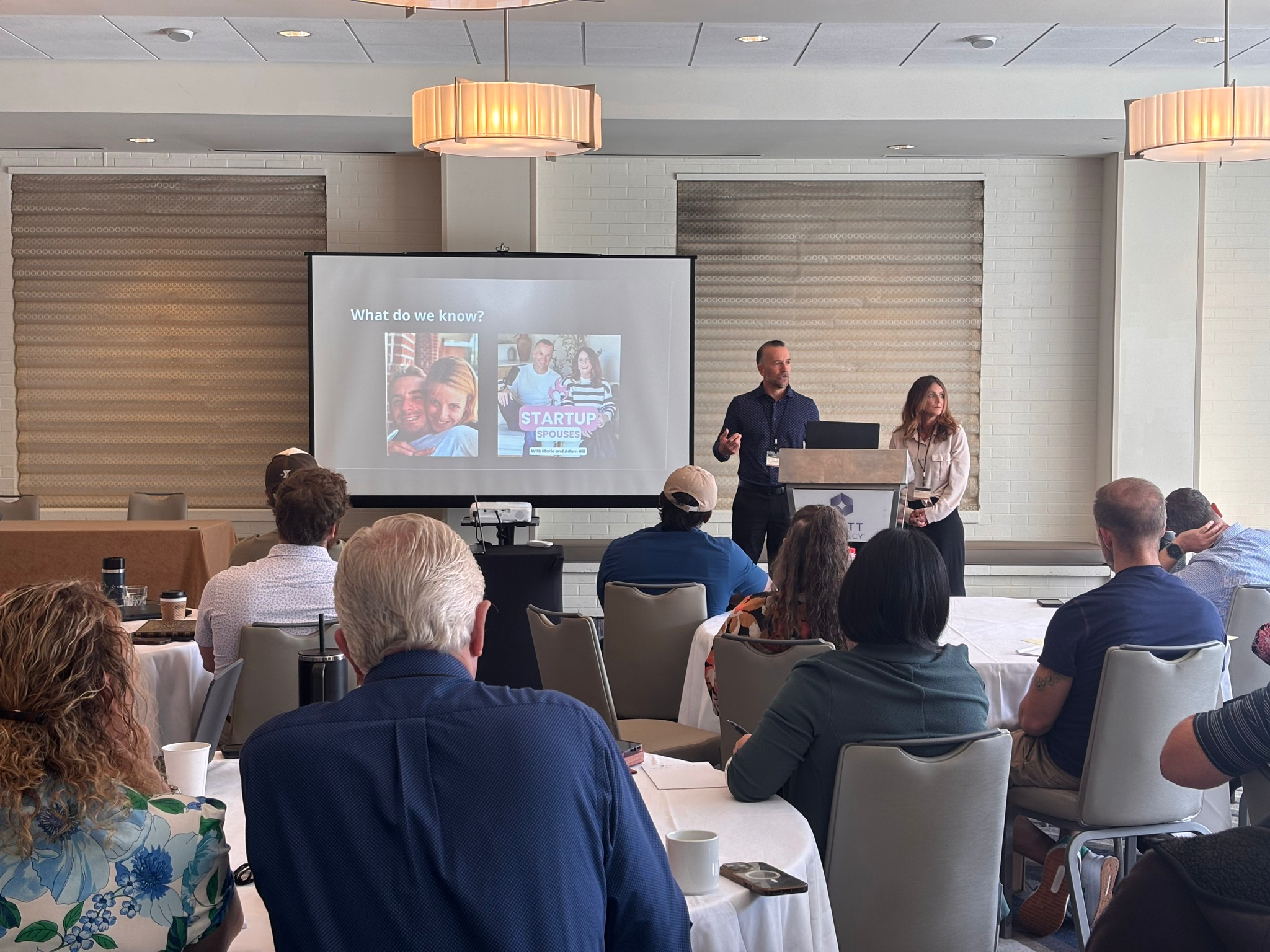 A conference or seminar room with a presentation screen displaying a slide titled 'What do we know?' with photos of a man and woman, and the title 'STARTUP SPOUSES.' Two presenters, a man and a woman, are standing at a podium with a laptop, addressing an audience seated at tables with white tablecloths and cups. The room has beige chairs, decorative window blinds, and ceiling lights.