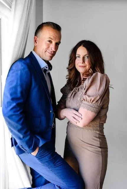 Adam Hill in a blue suit standing next to Marie Hill in a beige dress, both posing indoors near a window with white curtains.