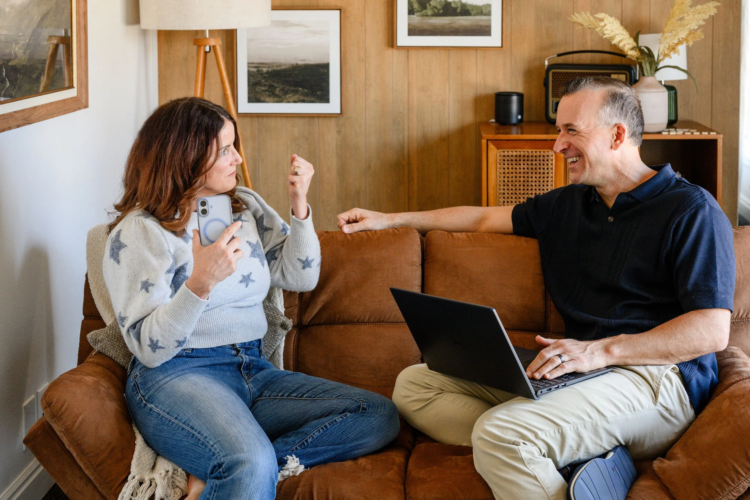 Marie Hill and Adam Hill sitting nearby with a laptop, smiling, teasing, and engaging in conversation inside a cozy, wood-paneled living room with framed pictures and decorative plants.