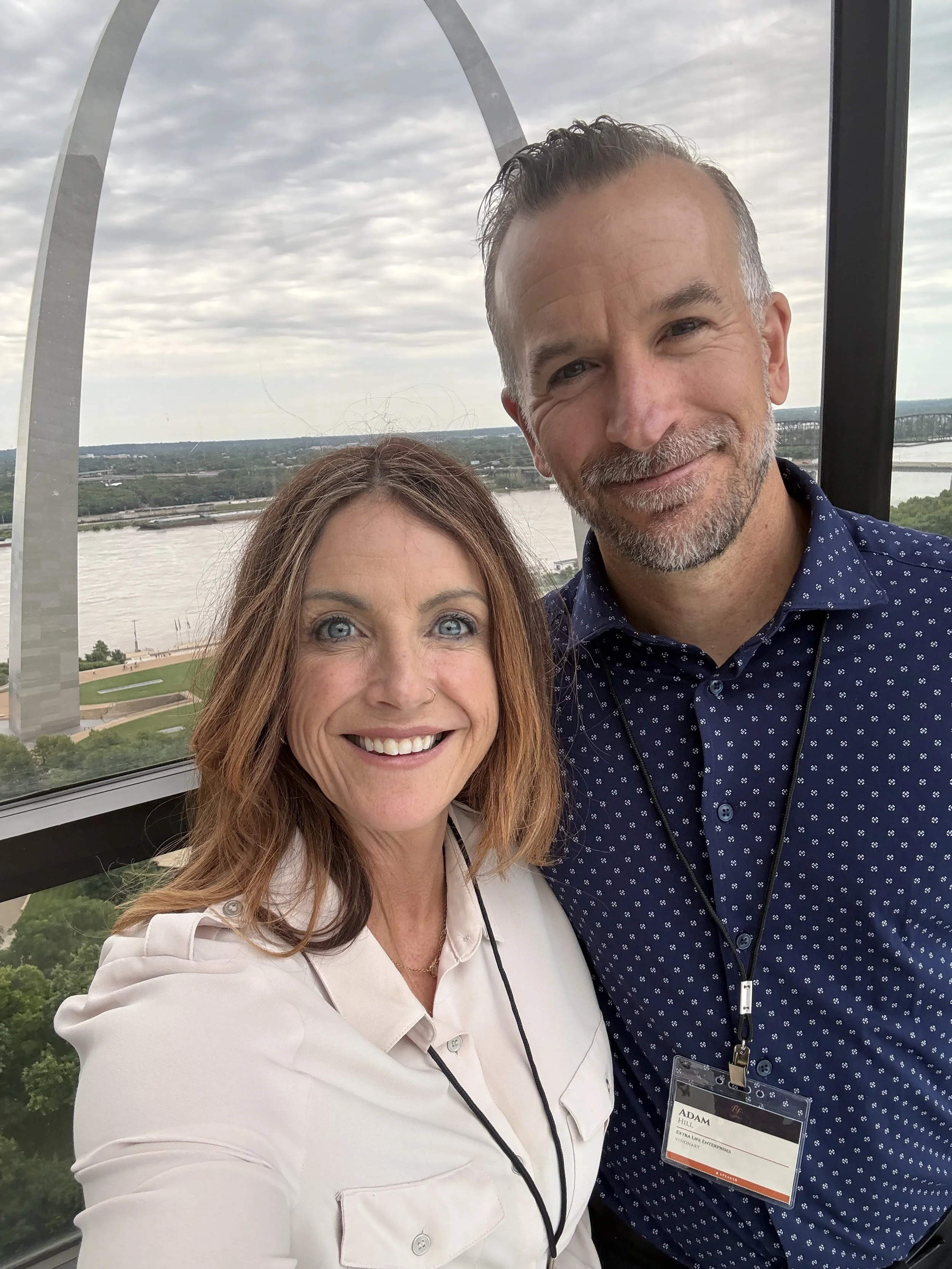 Close-up of Adam and Marie Hill taking a selfie in front of the Gateway Arch in St. Louis, Missouri, with a river and greenery in the background on a cloudy day.