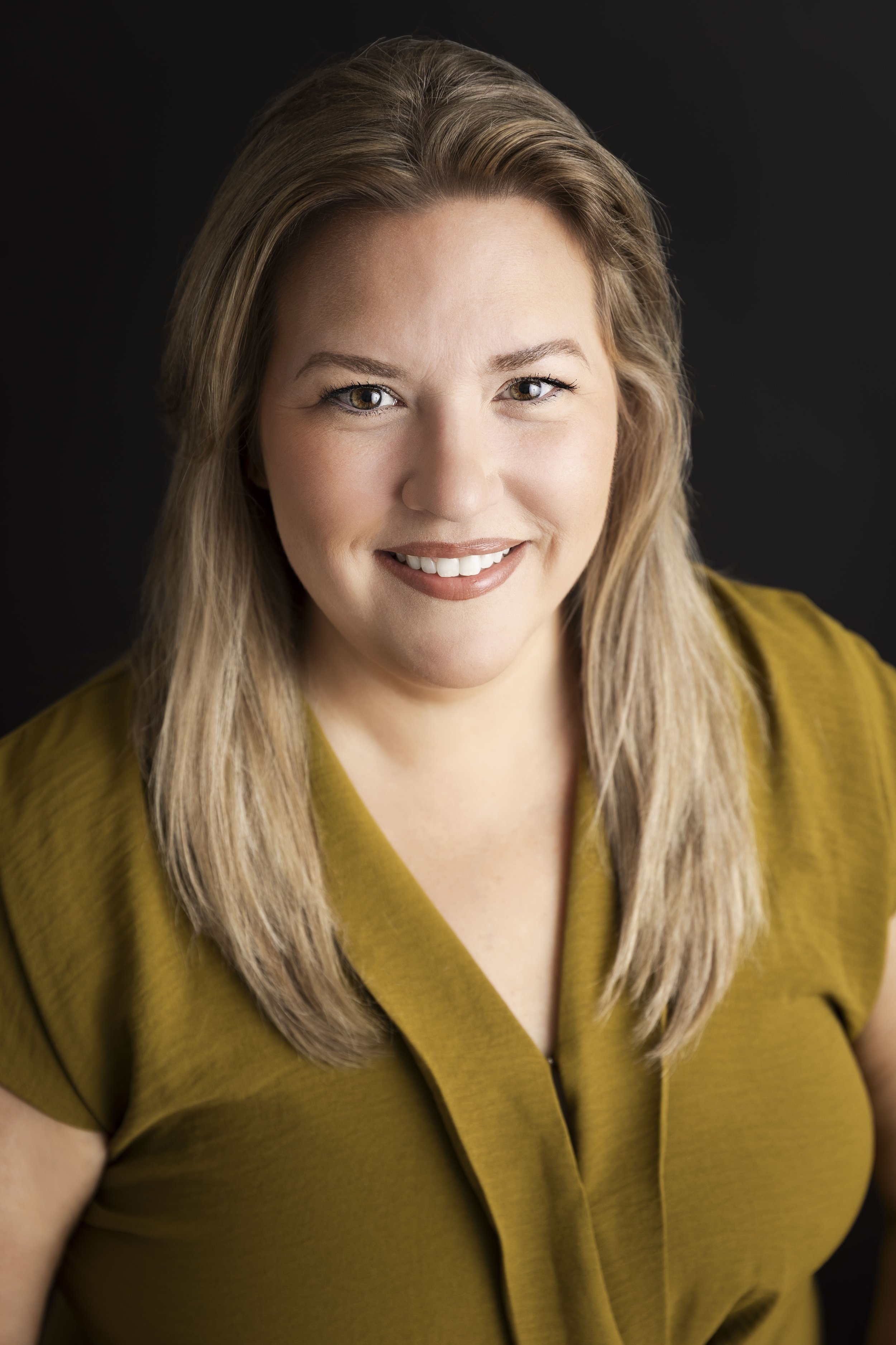 A woman with blonde hair, wearing a mustard-colored top, smiling against a black background.