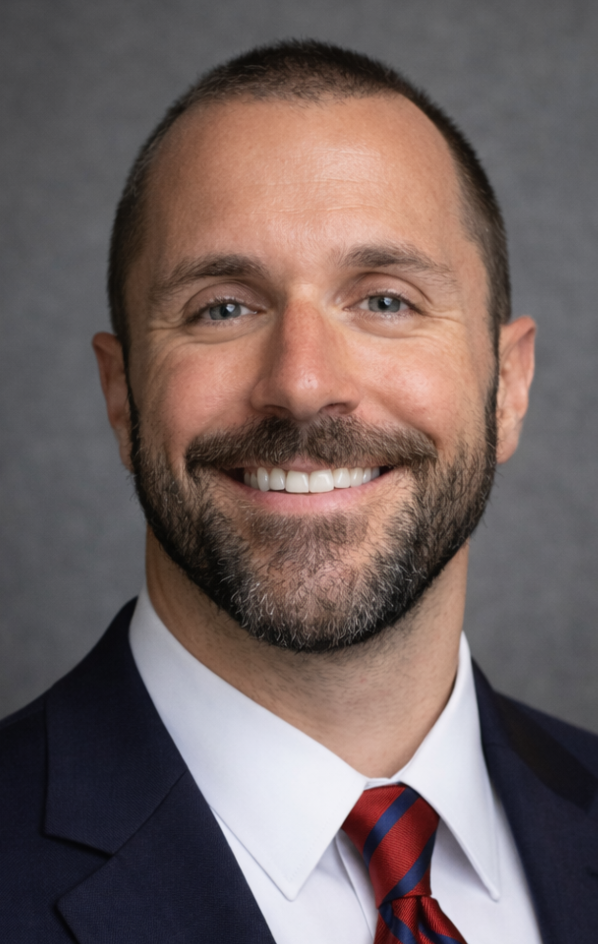 A smiling man with short dark hair, a beard, dressed in a dark suit, white shirt, and red striped tie, against a gray background.
