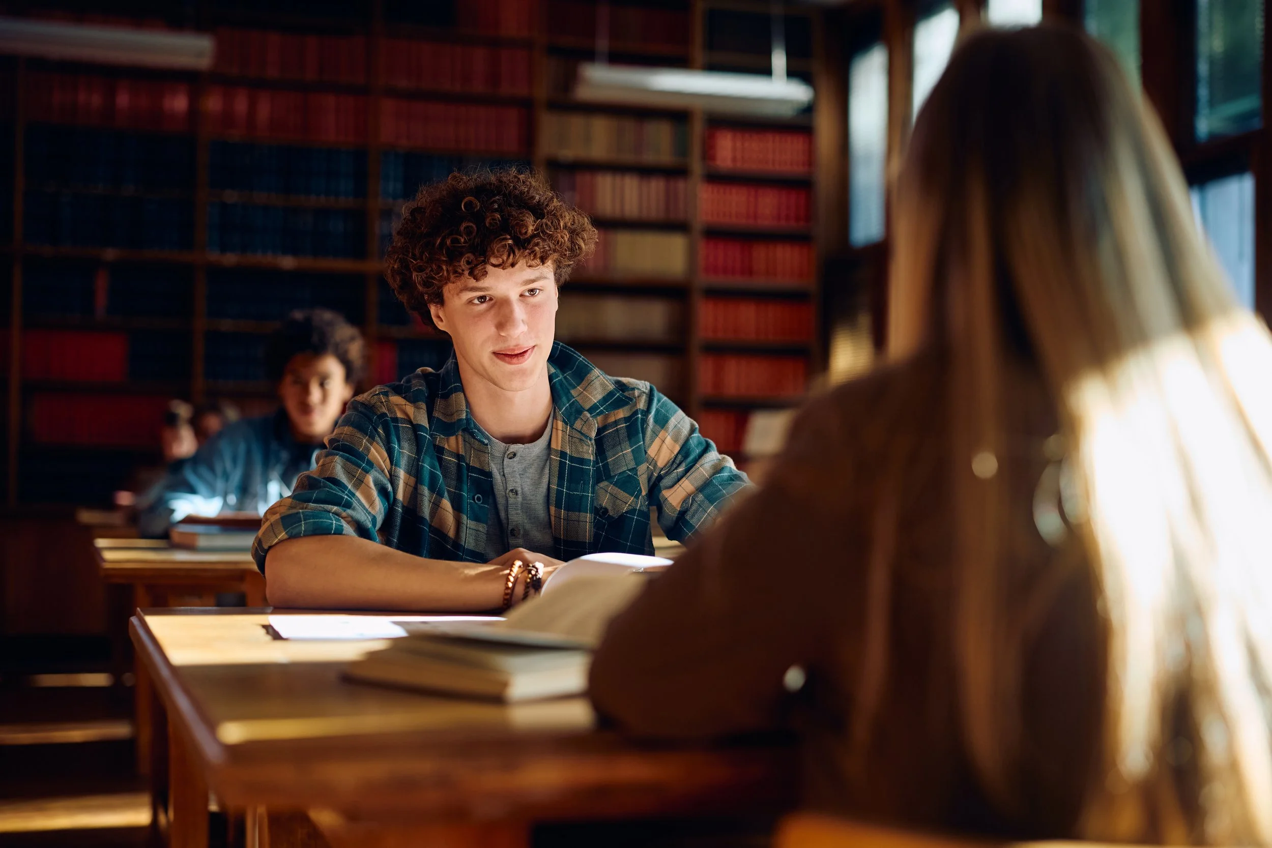 Teenager with curly hair wearing a plaid shirt talking to a girl with long blond hair in a library filled with books.