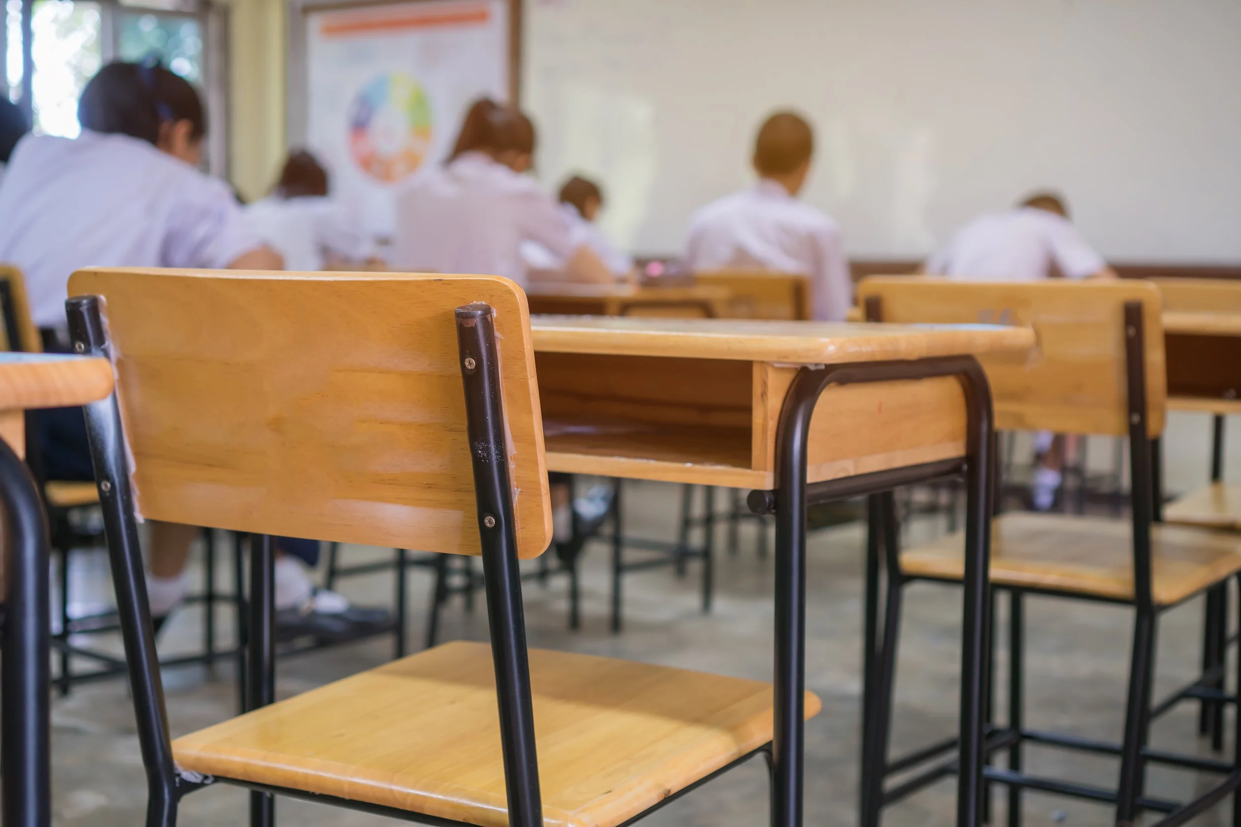 Empty classroom with wooden desks and students wearing white uniforms in the background.