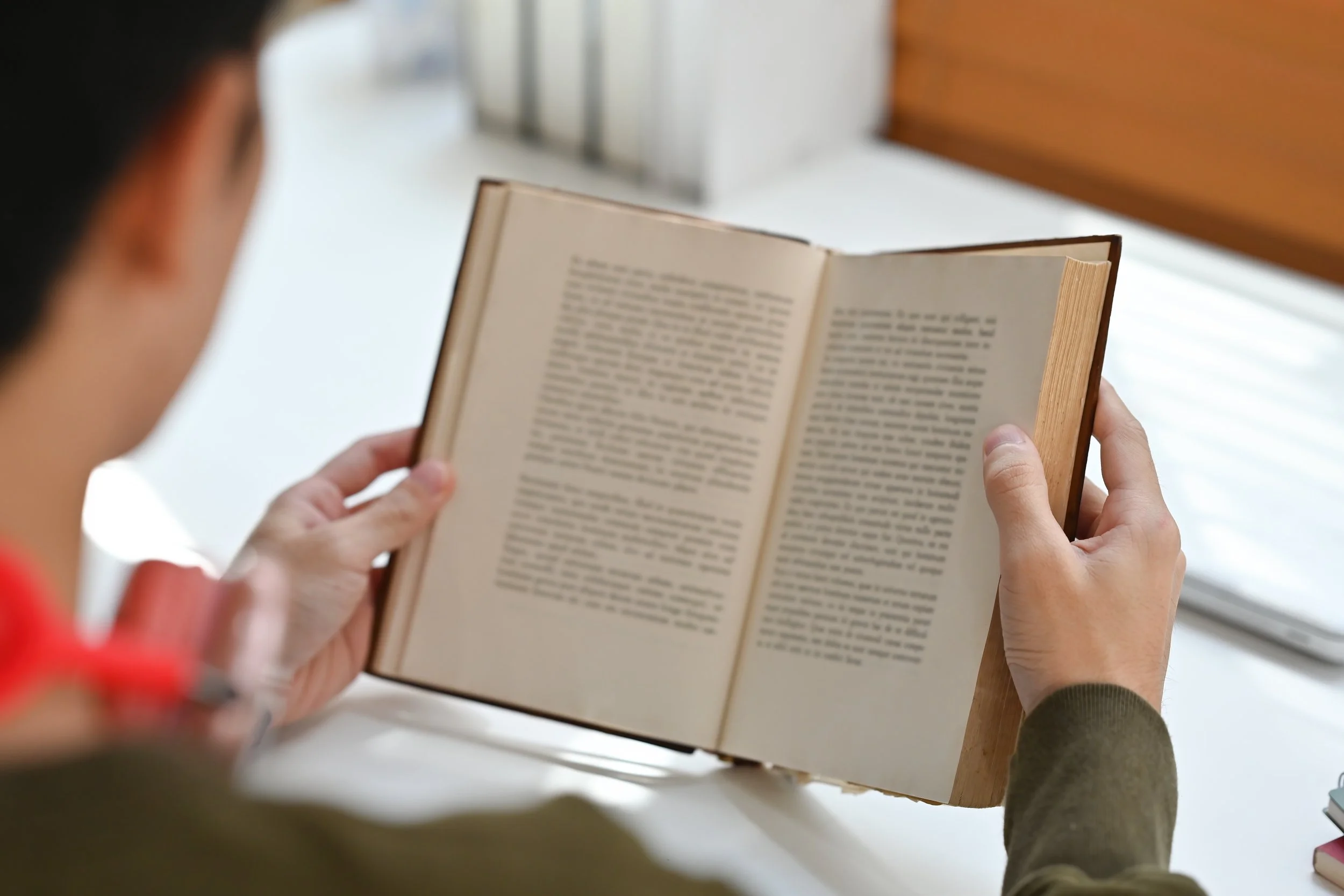 Person reading an open book with a blurred background of a window and desk.