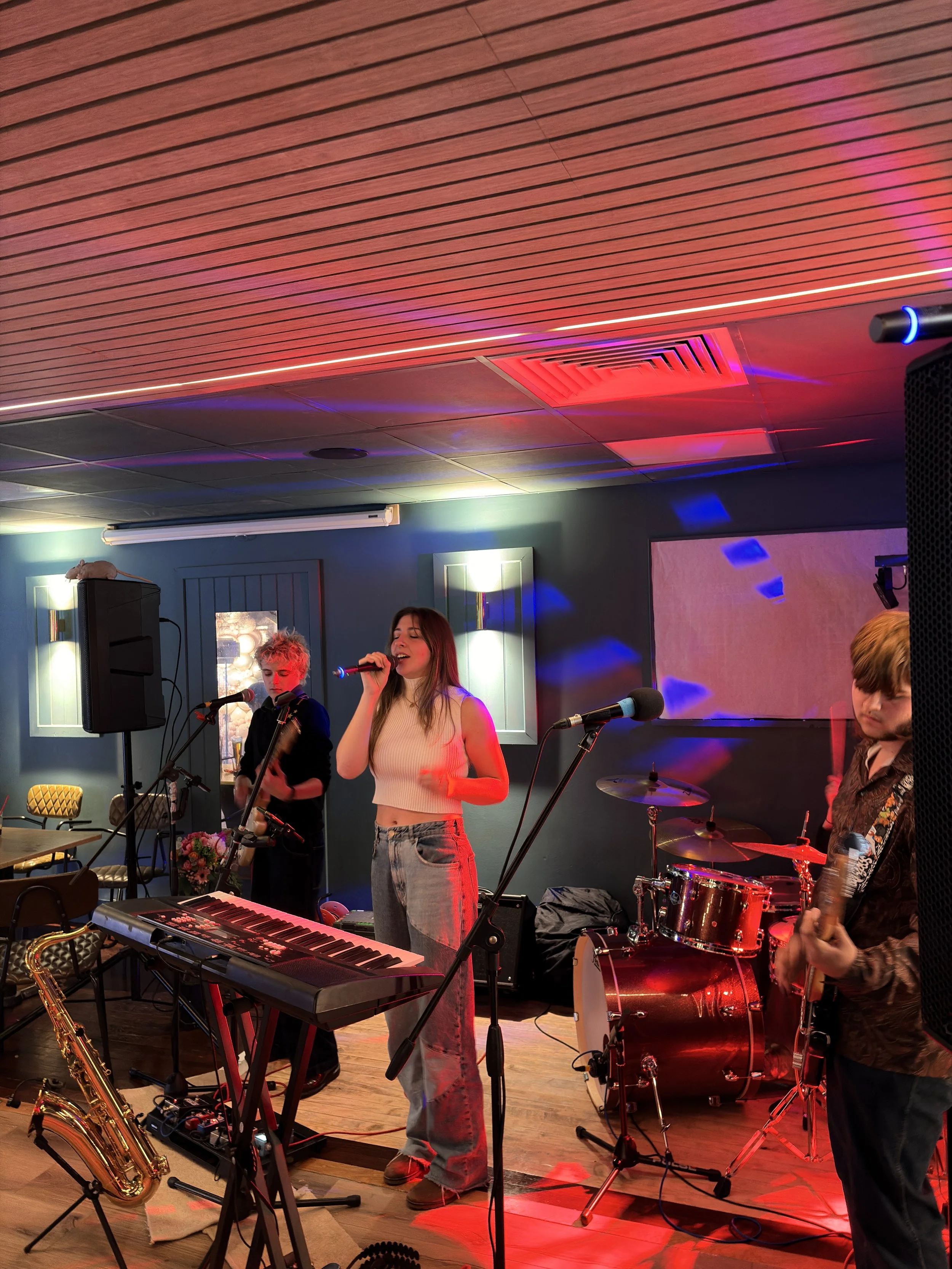 A band performing live on stage with a keyboard, drums, and guitar, in a cozy indoor venue illuminated with colorful stage lighting.