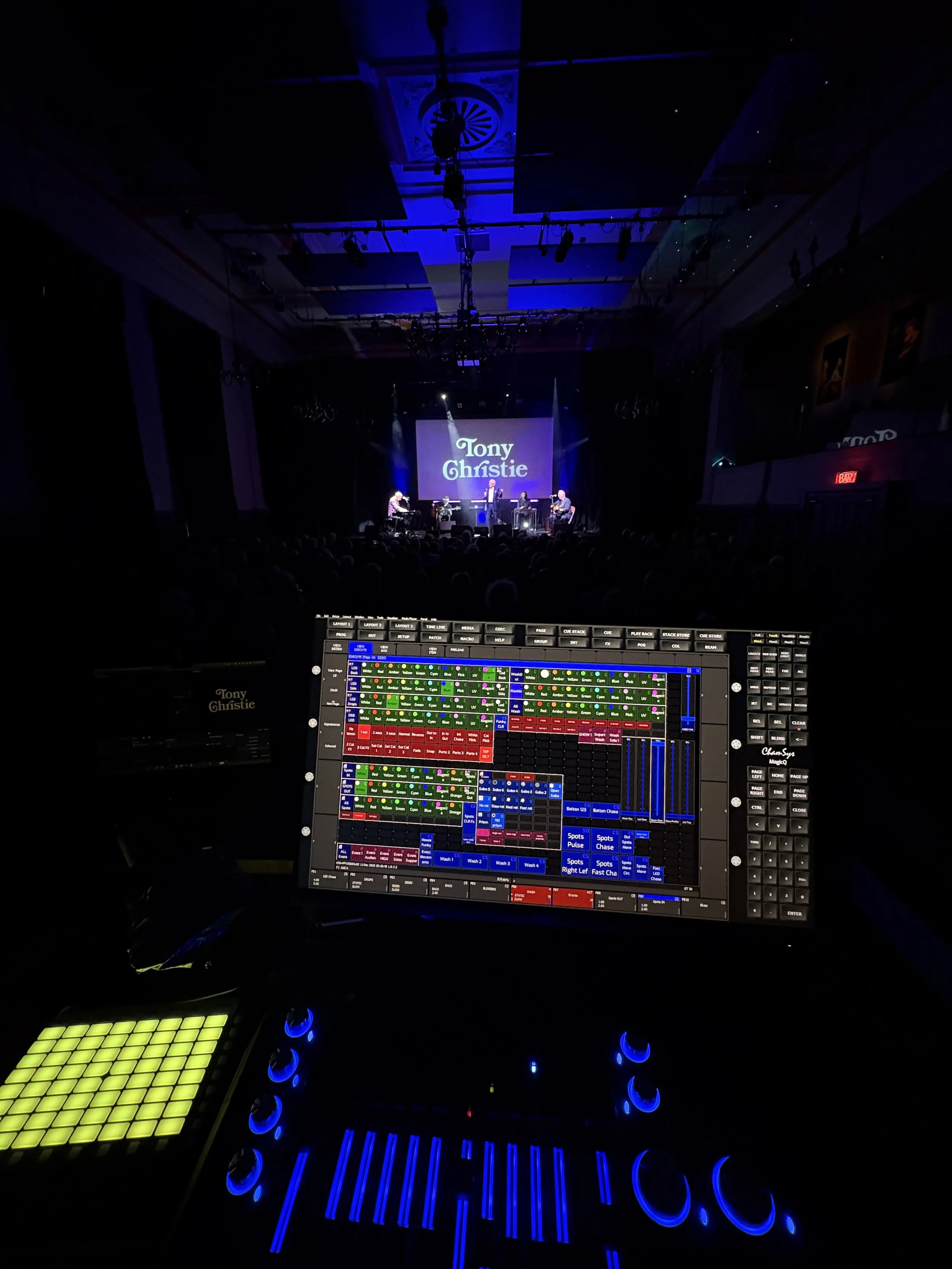 Stage with performers and a large screen displaying 'Tony Christie,' viewed from an audience with a control panel in the foreground.