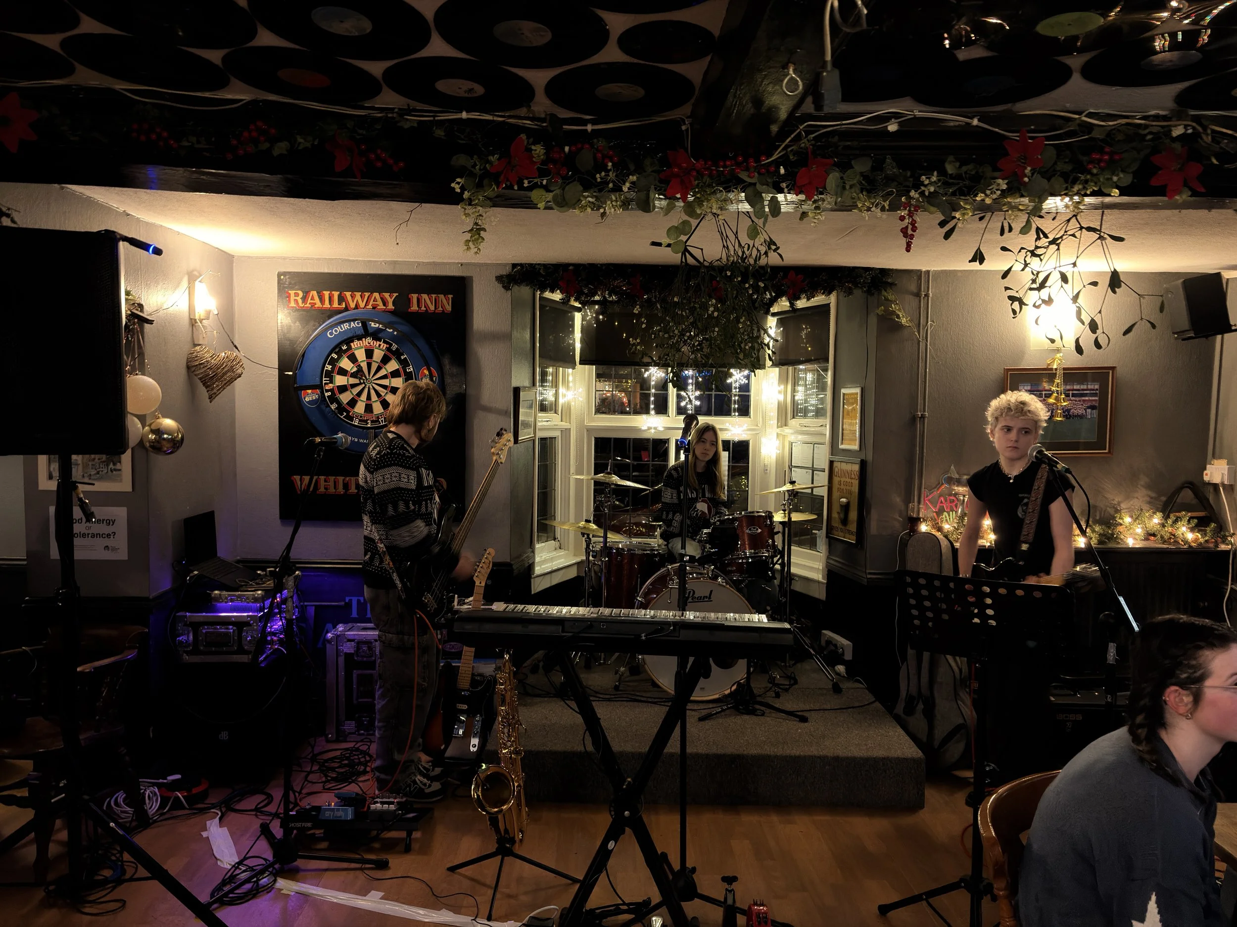 A small band performing on stage in a cozy pub. The band includes a drummer, a bassist, and a guitarist/vocalist. The pub has holiday decorations, including a string of lights, ornaments, and red poinsettias. There is a dartboard on the wall behind t