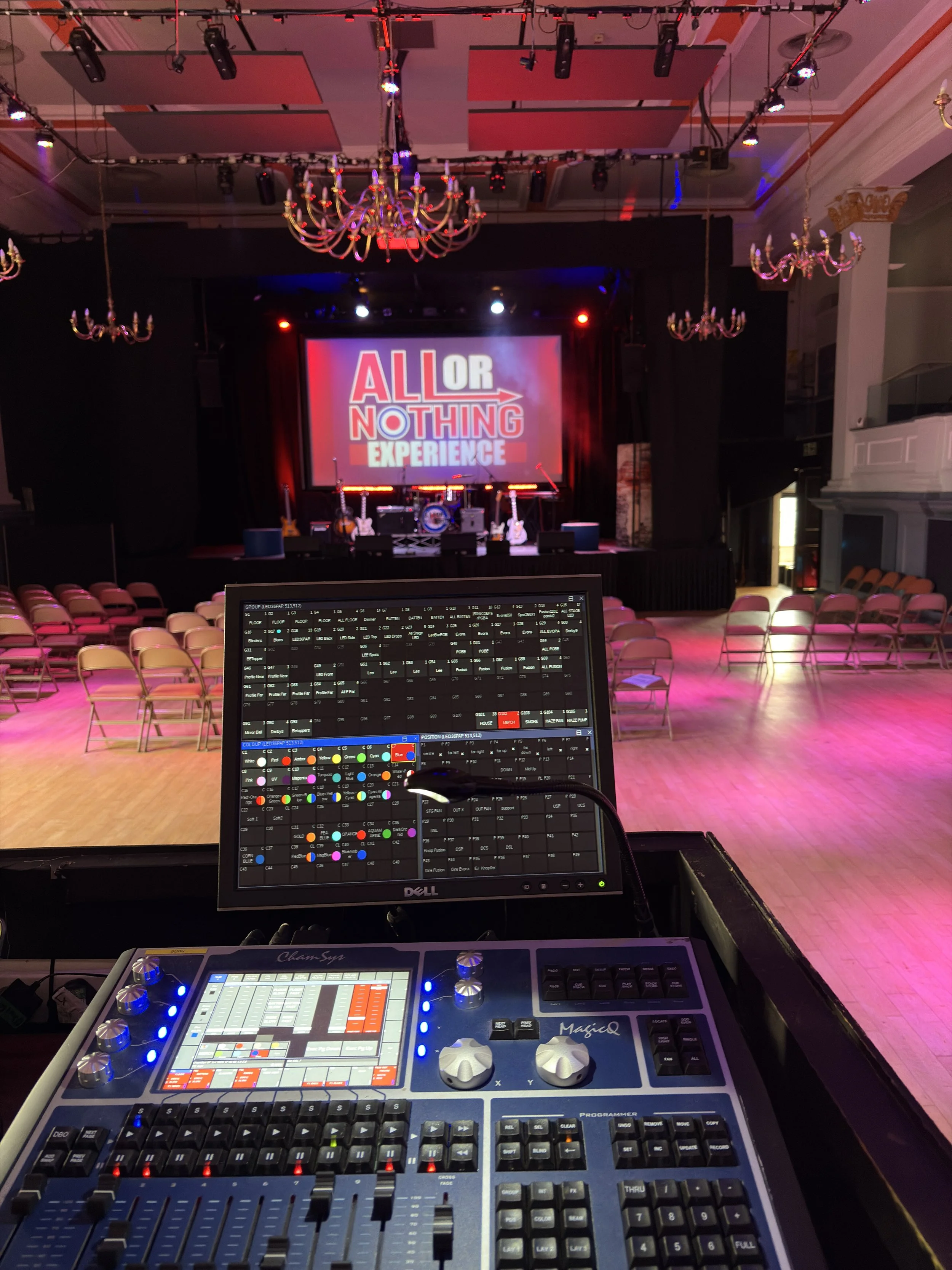 The image shows a stage with a large screen displaying the text "ALL OR NOTHING EXPERIENCE" in a music hall, with empty rows of chairs facing the stage. In front of the stage, there is a sound and lighting control console and computer monitor showing