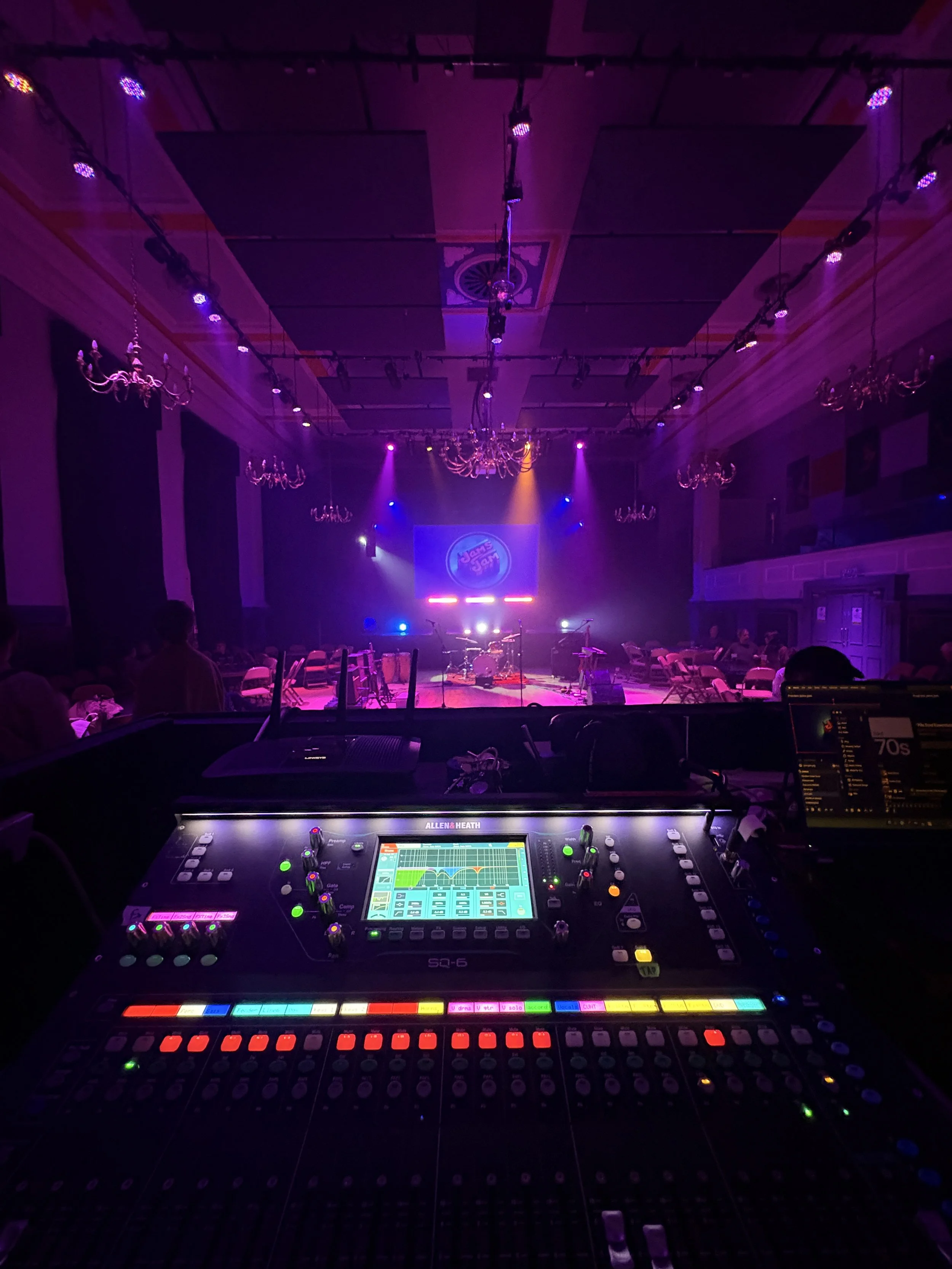 A stage with musical instruments under purple and blue lighting in a dimly lit concert hall. In the foreground, there is a mixing console with colorful buttons and screens.